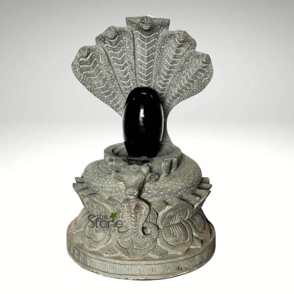 This image displays a highly detailed, sacred Hindu devotional object, meticulously carved from gray stone, depicting a Shiva Lingam with its associated iconography. At the center, a prominent Shiva Lingam stands, characterized by its smooth, highly polished, elongated oval form and a deep, reflective black color. This aniconic representation symbolizes the formless divine energy of Lord Shiva. The Lingam is nestled within a circular Yoni peetham (pedestal), also made of the textured gray stone, which is subtly designed with a small spout, typical for ritualistic ablutions such as abhishekam. Directly behind the Shiva Lingam, a magnificent five-headed cobra (Naga) rises, fanning out its intricately carved hoods to form a protective and majestic canopy. Each of the five cobra heads is individually sculpted with distinct eyes, mouths, and detailed scale patterns on their expanded hoods, emphasizing their mythological significance as protectors of the divine and as a symbol closely associated with Shiva. At the front of the multi-layered base, positioned with unwavering devotion, a serene, reclining Nandi bull statue faces the Shiva Lingam. Nandi, the sacred bull and Shiva's loyal mount, is rendered with noticeable horns, ears, and a calm, watchful expression. The circular pedestal supporting the Lingam and Nandi is further adorned with finely carved lotus petals and subtle textured patterns, possibly representing stylized coils or additional sacred motifs, symbolizing purity, spiritual awakening, and the cosmic order. The entire assembly rests upon a wider, stable, and slightly textured circular base, providing a solid foundation for this significant piece of religious art. The natural gray stone of the sculpture exhibits a granular, earthy finish, creating a beautiful textural and color contrast with the lustrous, smooth black Shiva Lingam. This intricate carving, combining the Shiva Lingam, Nagas, and Nandi, powerfully conveys themes of protection, devotion, and the cosmic energies central to Hindu worship, temple art, and spiritual practices. It serves as a potent visual representation of traditional Indian stone carving artistry and spiritual iconography.