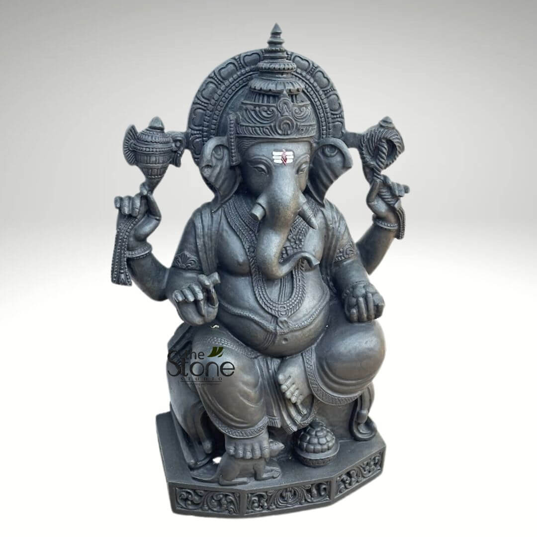 Ganapathi Statue for Home 2.5ft 2 striking, intricately carved dark grey stone sculpture of Lord Ganesha, appearing to be crafted from a dense material like granite or basalt, is depicted prominently against a clean, light grey gradient background that focuses all attention on the deity. The sculpture portrays the revered Hindu deity seated in the relaxed Lalitasana posture, often described as a posture of royal ease or comfort, with his right leg bent and foot resting flat on the ornate base, and his left leg folded across his lap. Ganesha, the elephant-headed god, is characterized by his large, fan-like ears, a single prominent tusk (symbolizing sacrifice and wisdom), and a long, gracefully curved trunk reaching towards a sweet modak (a traditional Indian dumpling) held in his lower left hand. A distinctive red and white Vaishnava tilak, a sacred mark representing spiritual devotion, adorns his forehead, set above serene, half-closed eyes that convey a sense of profound wisdom and tranquility. He possesses four arms, symbolizing his divine power and ability to interact with the world. His upper right hand holds a ceremonial axe (parashu), signifying detachment from worldly desires and the cutting away of ignorance. His upper left hand grasps a noose (pasha), representing the binding nature of earthly attachments or the ability to capture obstacles. His lower right hand rests open on his knee in the Varada Mudra, a benevolent gesture of granting blessings and boons to devotees. Ganesha's portly physique, a symbol of abundance, prosperity, and the fullness of creation, is adorned with finely sculpted necklaces, armlets, wristbands, and anklets, all rendered with meticulous detail. He wears a traditional dhoti-like lower garment with visible folds and patterns. A majestic, multi-tiered crown (mukuta) sits atop his head, complemented by an elaborately carved, semi-circular halo (prabhamandala) behind him, featuring intricate floral or geometric relief work. At his feet, a small, reverently carved mouse (Mushika), Ganesha's traditional vehicle or vahana, is depicted, embodying humility and the ability to overcome any obstacle, no matter how small. The entire sculpture rests on a robust, decorated pedestal, likely octagonal, featuring detailed relief patterns along its edges. The dark, monochromatic tone of the sculpture enhances its sculptural form and the depth of its intricate carving, with the even lighting highlighting every detail. A subtle watermark for "the Stone Studio" is visible in the lower center, indicating the craftsmanship and source. This devotional and artistic representation of Lord Ganesha, often worshipped as the "Remover of Obstacles," "Lord of Beginnings," and the deity of wisdom and prosperity, is perfect for articles discussing Hindu deities, spiritual practices, traditional Indian stone carving, religious art, cultural symbolism, meditation spaces, or home and garden decor focusing on auspicious themes and positive energy.