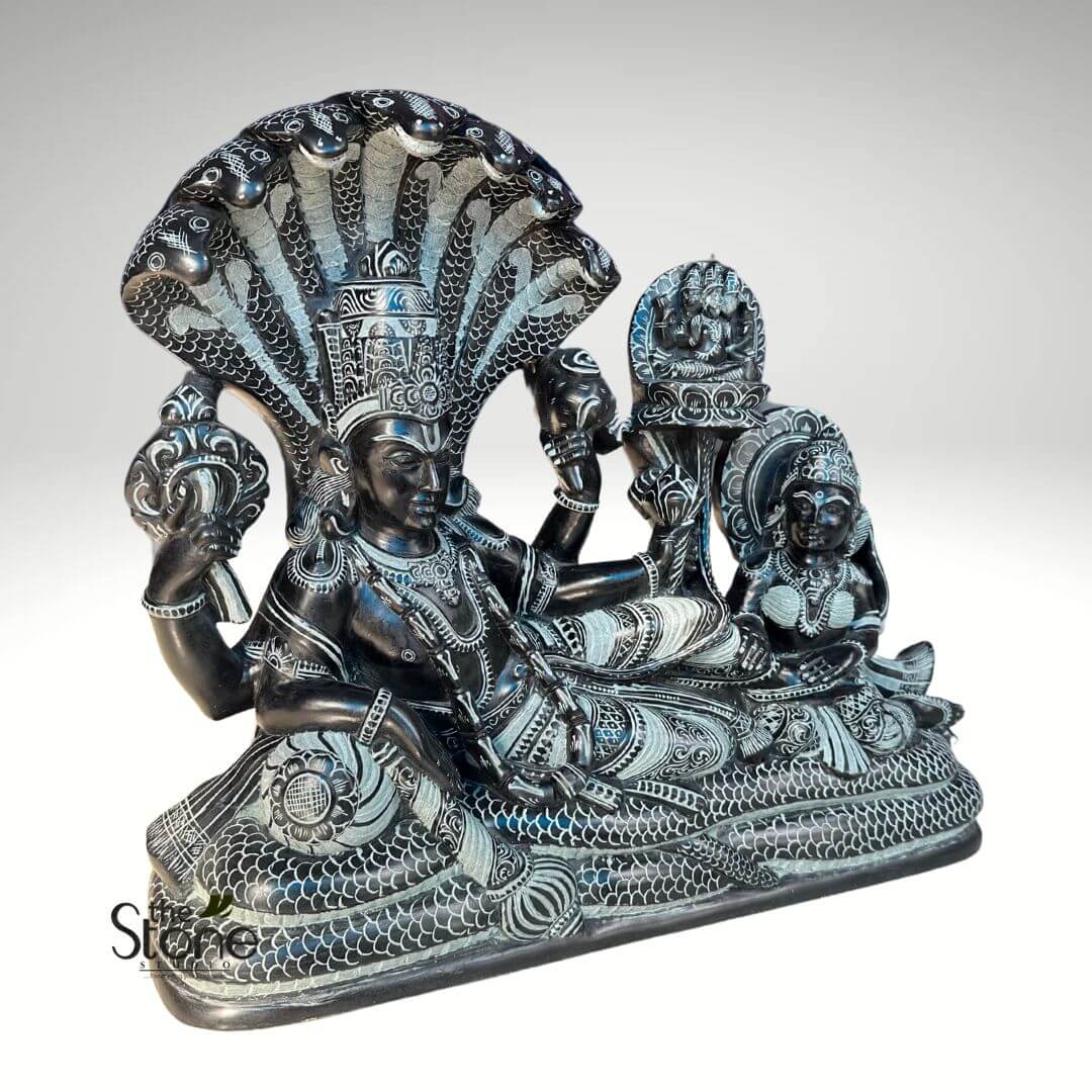 Lord Vishnu Sleeping on Sheshnag Statue 20" 2 magnificent and intricately carved dark stone sculpture depicts the Hindu deity Lord Vishnu in his iconic Ananthasayanam (cosmic slumber) pose, attended by Goddess Lakshmi. The entire sculpture is crafted from a dark, possibly black, stone, with lighter grey or white details meticulously carved into the surface, creating a striking contrast that highlights every ornate pattern and feature. In the center, Lord Vishnu, the preserver of the universe, reclines serenely on the coiled body of the multi-headed serpent Ananta (also known as Sheshnag). Vishnu's body is richly adorned with elaborate necklaces, armlets, bracelets, and a finely patterned dhoti, all rendered with incredible detail. He possesses four arms; his upper right hand holds the Sudarshana Chakra (discus), and his upper left hand grasps the Panchajanya Shankha (conch shell). His lower right arm rests gracefully on his side, while his lower left arm extends towards Lakshmi. His face bears a calm, meditative expression, with eyes half-closed, and he wears an ornate, towering crown (kiritamukuta) that is also intricately carved. Behind Vishnu's head, the colossal serpent Sheshnag forms a majestic canopy with its seven or nine fanned-out cobra heads, each head detailed with scales and patterns. Vishnu's entire body rests upon Sheshnag's massive, coiled form, which is meticulously sculpted with a textured scale pattern along its length, creating a foundational bed for the deity. At Vishnu's feet, to the right of the image, Goddess Lakshmi, the deity of wealth and prosperity, is seated in a pose of devotion and attendance. She is beautifully adorned with jewelry and garments that echo the intricate carving style of Vishnu. Her expression is attentive, and her gaze is directed towards her consort, reinforcing the scene of divine cosmic repose and reverence. The entire mythological tableau rests on a simple, flat, rectangular base, elevating the figures. This exquisite dark stone carving, with its contrasting light details, is a prime example of traditional Hindu iconography and sculpture, symbolizing cosmic order, divine preservation, and the eternal cycle of time. It would be highly relevant to an article discussing Hindu deities, religious art, ancient Indian craftsmanship, or the symbolism of Vishnu's cosmic slumber.