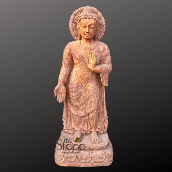 majestic, full-length standing Buddha statue, intricately carved from reddish-brown natural stone, likely sandstone or a similar earthy-toned material, is presented against a plain, dark gray background. The sculpture showcases a finely textured, slightly weathered appearance, highlighting the skilled craftsmanship. The Buddha's face is rendered with a profound sense of serenity and inner peace, featuring downcast, contemplative eyes, a gentle smile, elongated earlobes reaching almost to his shoulders, and a prominent urna (a dot or tuft of hair between the eyebrows) symbolizing spiritual insight. His hair is meticulously carved in tight, small curls, culminating in an ushnisha (a cranial bump) at the top of his head, denoting wisdom and enlightenment. Behind his head, a beautifully detailed circular aureole or halo suggests his divine status, possibly depicting stylized flames or lotus petal motifs, adding to the deity's sacred aura. His posture is upright and graceful, with the right hand raised to shoulder height in the Abhaya Mudra, a gesture of fearlessness, blessing, and protection, with the palm facing outwards and fingers extended. The left hand is lowered and extended outwards, possibly in the Varada Mudra (granting wishes) or a similar gesture of generosity and compassion, inviting contemplation and spiritual connection. The Buddha is draped in a flowing, finely sculpted monastic robe that accentuates the contours of his body with naturalistic, cascading folds, particularly visible across his chest and down his legs, highlighting the artisan's mastery in stone carving and drapery. The robe's texture and folds add depth and realism to the figure. The figure stands barefoot on an elaborate, multi-tiered lotus pedestal, a classic symbol of purity, beauty, and spiritual awakening in Buddhist iconography. The base itself is carved with intricate details, complementing the overall elegance of the statue. The entire sculpture is presented against a plain, dark gray to black background, which serves to dramatically emphasize the statue's warm, earthy tones and intricate details, making it the sole focal point. The soft, even lighting enhances the dimensionality of the relief carvings and the subtle textures of the stone. This exquisite piece exemplifies traditional Buddhist art and sculpture, demonstrating exceptional craftsmanship and a deep understanding of spiritual iconography. It would be an ideal focal point for discussions on spiritual décor, the history of stone carving, Asian art, or the aesthetic appeal of religious artifacts for spaces seeking a sense of peace and meditation. The subtle "the Stone studio" watermark at the bottom left suggests its origin or craftsmanship, linking it to themes of artisan studios, custom stone work, or sourcing high-quality spiritual art pieces.