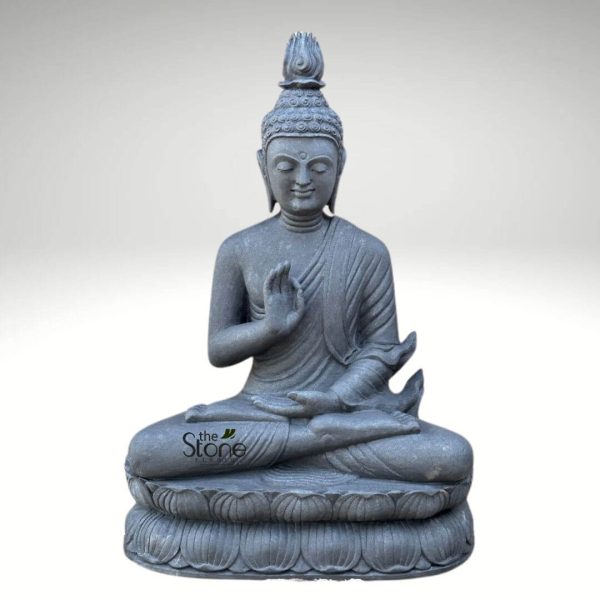 detailed and serene monochromatic gray stone statue of Buddha, carved from what appears to be dark granite or basalt, stands prominently against a clean, light gray background. The Buddha is depicted in a full meditative pose, seated gracefully in the traditional cross-legged Padmasana (lotus position), symbolizing stability, deep contemplation, and spiritual awakening. His right hand is raised in the Abhaya Mudra, a gesture of fearlessness, reassurance, and divine protection, with the palm facing outwards and fingers gently extended upwards. His left hand rests softly in his lap, palm facing upwards in a gesture often associated with meditation (Dhyana Mudra), symbolizing openness and receptivity to spiritual wisdom. Buddha's face is characterized by profound tranquility and inner peace, with eyes gently closed or cast downwards in introspection. A faint, serene smile graces his lips, and his elongated earlobes, symbolic of his princely past and renunciation of worldly possessions, are clearly visible. His head is adorned with tightly coiled curls of hair, culminating in a distinctive flame-like Ushnisha at the crown, which signifies his wisdom, spiritual enlightenment, and expansive consciousness. The monastic robes are elegantly draped over his left shoulder, leaving the right shoulder bare, and are rendered with natural, flowing folds and pleats that convey the texture and movement of fabric, despite being carved in solid stone. The entire figure rests upon an intricately carved double-lotus pedestal, a sacred and symbolic base, with each petal distinctly sculpted to add depth and detail to the foundation. The matte, textured finish of the stone enhances the statue's antique and natural aesthetic, highlighting the artisan's craftsmanship. A subtle watermark "the Stone Studio" is visible in the lower left section, identifying the creator or vendor of this magnificent piece. This handcrafted Buddha sculpture is presented as a spiritual art object, ideal for creating a tranquil atmosphere in meditation spaces, spiritual decor settings, or as an elegant garden focal point, connecting directly to themes of mindfulness, peace, and timeless artistry often explored in articles about such artifacts.