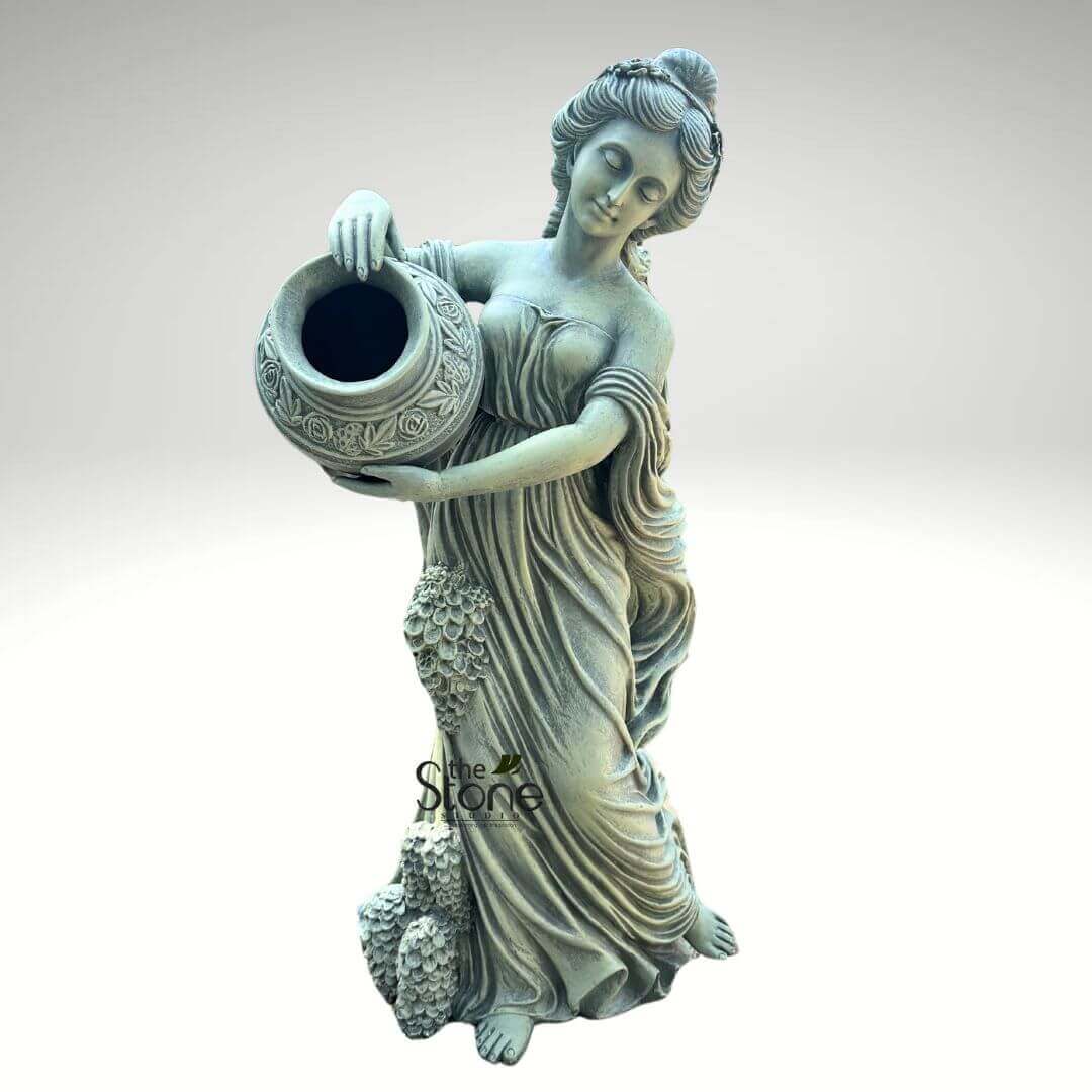 Water Fountain Lady Statue 5ft 6 full-body, classical-style garden statue crafted from light grey, weathered stone or concrete, depicting a graceful female figure in a tranquil, contemplative pose. The woman's head is gently tilted downwards, with her eyes softly closed, conveying a sense of serenity. Her hair is intricately styled in an updo, adorned with what appears to be a delicate headband or wreath. She is draped in flowing, Hellenistic-style robes that softly gather around her bust and waist, cascading in elegant folds to her bare feet. She holds a large, ornate urn or amphora with both hands, positioned at her hip level as if presenting or pouring from it. The vessel features finely detailed, raised relief patterns of floral and vine motifs, adding to its classical charm. Its dark, open mouth suggests it could function as a planter for flowers or greenery, or potentially as part of a subtle water feature in a garden setting. A distinctive and prominent feature of the statue is a lavish cluster of textured grapevines and bunches of grapes that gracefully spill down the left side of her gown, from just below her waistline to her feet. This element strongly evokes themes of harvest, abundance, and ancient mythological figures like Dionysus or Bacchus. The stone material exhibits a slightly aged, matte finish with subtle greenish undertones in certain recesses, enhancing its antique appeal and indicating its suitability for durable outdoor display. A small, subtle watermark logo for "The Stone" is visible near the base of the statue, indicating its origin or craftsmanship. This detailed piece is ideal for enhancing sophisticated outdoor spaces, classical gardens, or vineyard-themed landscapes with a touch of timeless elegance and artisan stone sculpture.