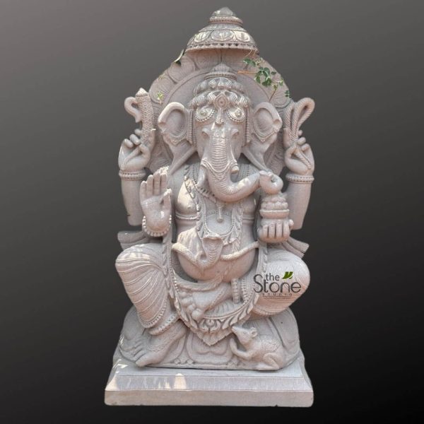 highly detailed, meticulously carved stone statue of Lord Ganesha, fashioned from light-colored, possibly white or grey, natural stone, stands prominently against a plain, dark grey background. The elephant-headed Hindu deity is depicted seated in a relaxed yet regal posture, likely a variation of Lalitasana, on a simple rectangular plinth. His prominent potbelly, symbolizing prosperity and abundance, is visible beneath his richly adorned chest. Ganesha's benevolent face features large, intricately carved ears, which fan out gracefully, emphasizing his ability to listen to devotees' prayers. He wears an elaborate crown (mukut) adorned with delicate patterns. His trunk curves gently towards his lower right hand, reaching for a bowl filled with modaks (sweet dumplings), a treat he is famously fond of. Notably, one of his two tusks is broken, a characteristic often associated with his role as a scribe of the Mahabharata. The deity is depicted with four arms, each holding a symbolic object. His upper right hand grasps an ankusha (elephant goad), representing control over obstacles, while his upper left hand holds a pasha (noose), symbolizing restraint from worldly attachments. His lower left hand is raised in the Abhaya mudra, a gesture of blessing, reassurance, and fearlessness, offering protection to his devotees. Ganesha is lavishly adorned with multiple necklaces, intricate armlets, bracelets, and anklets, all meticulously sculpted from the same stone. The drapery around his lower body shows elegant folds and patterns, suggesting fine, traditional clothing. At his feet, to the right, sits his loyal vahana (mount), a small, intricately sculpted mouse (Mushika), looking up at him, symbolizing Ganesha's ability to overcome even the smallest obstacles. Behind Ganesha is an elaborate prabhavali (decorative arch or aura) featuring delicate floral and scrollwork designs, adding to the grandeur of the statue. The top of the prabhavali is crowned with a distinctive finial, and faint hints of green, possibly painted details or slight organic growth, are visible on its upper curves. This exquisite piece of spiritual art showcases remarkable artisan craftsmanship, highlighting the intricate details possible with traditional Indian stone carving techniques. The statue embodies prosperity, wisdom, and the removal of obstacles (Vighnaharta), making it a significant representation of Hindu faith and culture. A subtle "the Stone Studio" watermark is visible in the lower right portion, indicating its origin. This detailed portrayal connects deeply to articles discussing Hindu religious art, traditional Indian sculpture, spiritual home decor, the significance of Lord Ganesha, or the mastery of stone carving in India.