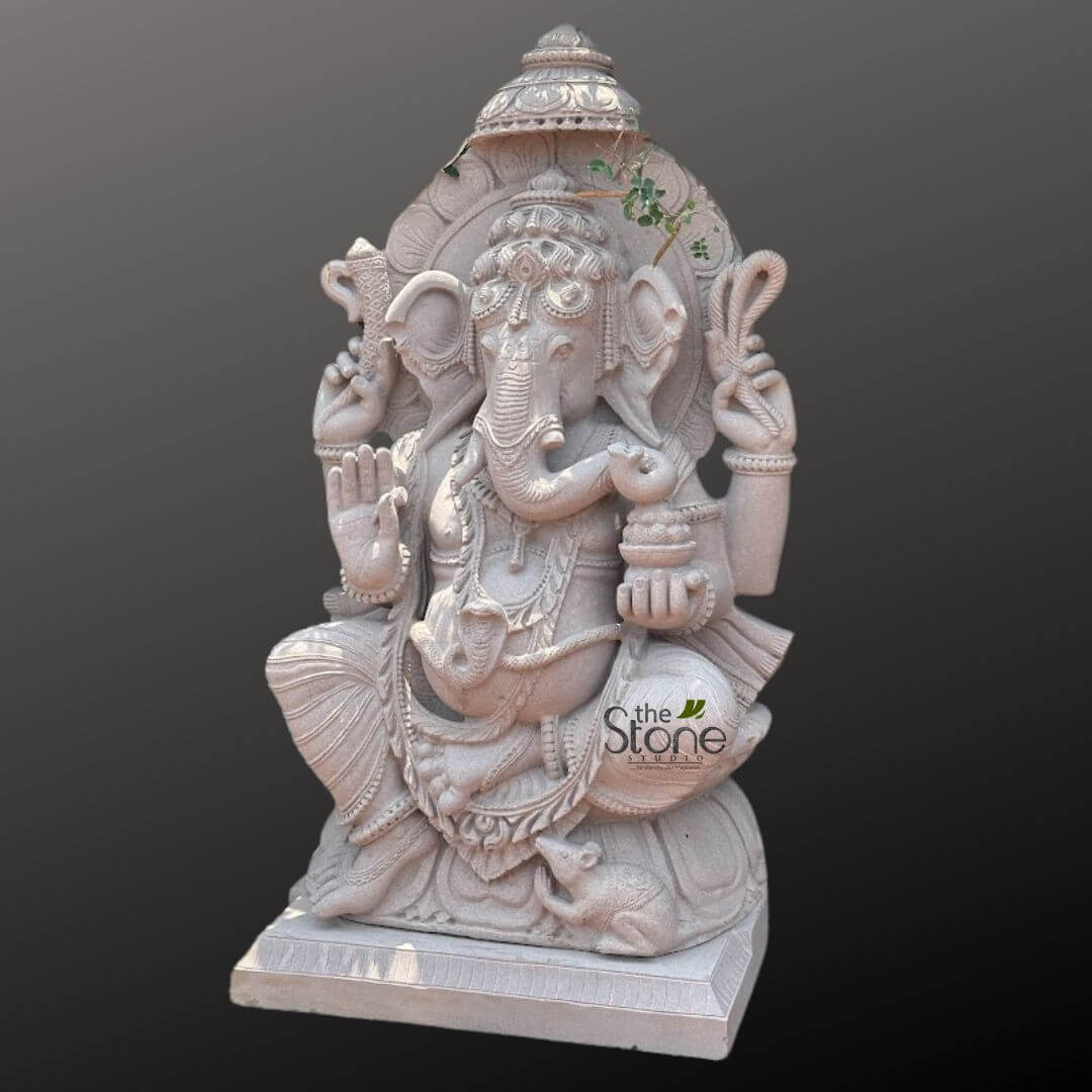Big Ganesha Idol for Outdoor 4ft 4 magnificent, finely carved stone statue of Lord Ganesha, presented in a striking light grey or off-white hue, suggesting the use of high-quality marble or sandstone. The Hindu deity is depicted seated in a relaxed yet regal posture, likely Lalitasana, with one leg folded and the other slightly raised, conveying an air of benevolent wisdom and approachability. His iconic elephant head features prominent, fanned ears and a gracefully curved trunk, which subtly reaches towards the offering in his lower left hand. A single, distinct tusk (symbolizing wisdom and sacrifice) is visible. He is adorned with an elaborate, multi-tiered crown and intricate jewelry, including earrings and a forehead ornament, all carved with exceptional detail that highlights the artistry of Indian stone carving. With four arms, Lord Ganesha holds his traditional attributes: the ankusha (goad) in his upper right hand, and the pasha (noose) in his upper left, symbolizing control over worldly desires and attachments. His lower right hand is raised in Abhaya Mudra, offering blessings and dispelling fear. In his lower left hand, he cradles a bowl overflowing with modaks or ladoos, his favorite sweet treats, signifying prosperity and abundance. His prominent, pot-bellied form, symbolizing wealth and generosity, is draped in a meticulously carved dhoti with elegant pleats and patterned borders. He wears multiple necklaces, armlets, and bracelets, along with a sacred thread (upavita) across his chest, all testament to the handcrafted artistry. At his feet, his faithful vahana, the mouse (Mooshika), is depicted, adding an authentic touch to the religious idol. The entire stone sculpture rests on a sturdy, multi-tiered rectangular pedestal, designed to elevate and showcase the deity. Behind Ganesha, an ornate, semi-circular prabhamandala or aura is carved, featuring lotus petal designs and a decorative finial at the top. Notably, a small sprig of green foliage, perhaps a vine, has naturally grown into a crevice of the prabha, hinting at its potential placement in a garden or outdoor setting, or simply adding a touch of natural beauty. The image is set against a plain, solid dark grey background, which serves to isolate and highlight the intricate details and texture of the Ganesha statue. A subtle watermark for 'the Stone Studio' is visible on the lower right, identifying the source of this exquisite piece of traditional Indian stone art. This Ganesha statue exemplifies premium craftsmanship and would be a significant addition to any home altar, spiritual space, or as a focal point in garden decor, symbolizing good fortune, wisdom, and the removal of obstacles for those seeking a majestic and spiritually significant stone sculpture.
