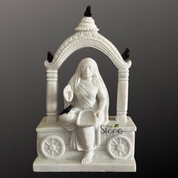 highly detailed, pristine white marble sculpture depicts a serene female deity or saint seated gracefully within an ornate, miniature shrine. The figure is draped in flowing, intricately carved traditional robes that cascade around her form, revealing bare feet resting on the base. Her long, sculpted hair flows over her shoulders. Her right hand is raised in the Abhaya Mudra, a gesture of fearlessness and blessing, while her left hand gently holds a small, rectangular basket or tray, possibly for offerings. She is framed by an ornate archway, supported by two sturdy, fluted pillars. The arch is richly adorned with delicate, traditional Hindu patterns, including a row of beads and a serrated edge, topped with a small finial. Contrasting dramatically with the luminous white marble, four small, dark black crows are strategically perched on the sculpture: one sits atop the finial, two rest on the curves of the archway, and one is near the figure's left hip, close to the basket. These dark birds add a significant symbolic layer, often associated with spiritual wisdom, messengers, or specific deities like Shani or a compassionate form of Parvati (such as Annapurna, known for providing sustenance) in Hindu mythology. The entire shrine rests on a stylized rectangular base, which features two visible, spoked wheels on each side, suggesting a mobile temple or a processional chariot (ratha). The craftsmanship highlights the natural purity and luminosity of the marble, creating a tranquil and devotional artifact. The background is a plain, dark grey, emphasizing the intricate details and smooth finish of the white marble sculpture. This detailed piece serves as an exemplary representation of traditional Indian spiritual art, embodying themes of sustenance, nature, divine blessing, and protection, making it a focal point for an article on Hindu iconography, marble craftsmanship, or cultural symbolism. A "The Stone Studio" watermark is visible on the lower right of the base.