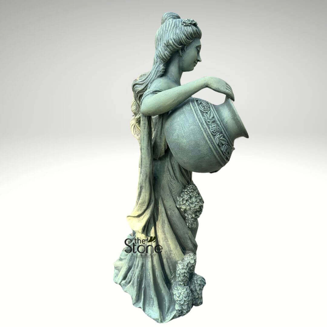 Water Fountain Lady Statue 5ft 5 detailed, classical-style outdoor garden statue of a serene maiden or goddess is captured in a graceful side profile against a plain, light grey background. This large, elegant sculpture, likely crafted from durable concrete or natural stone, exhibits a weathered, antique finish in a muted grey-green tone, suggesting age and timeless beauty. The figure stands with her head gently bowed, looking down at a substantial, ovoid urn or water pot that she cradles in her arms, poised as if to pour. Her elegant coiffure features intricately carved flowing locks and an updo, adorned with a delicate floral piece above her ear. She is draped in classical, flowing attire, characterized by soft folds and gathers that define her form, reminiscent of ancient Greek or Roman garments. Adorning her lower dress are clusters of meticulously sculpted grapes and foliage, adding to the natural, bountiful theme. The vessel itself is a work of art, featuring a richly textured, raised band of decorative floral and vine patterns around its upper half, complementing the statue's classical aesthetic. The craftsmanship highlights fine details in the drapery, hair, and the decorative elements on both the figure and the pot. This versatile piece is ideal for landscape design, serving as a focal point in a garden, patio, or as part of a tranquil water fountain or pond feature. Its durable material and elegant design make it perfect for enhancing outdoor living spaces with a touch of traditional artistry. A small "the Stone Studio" watermark in the bottom left corner indicates its origin as a high-quality product for home and garden decor.