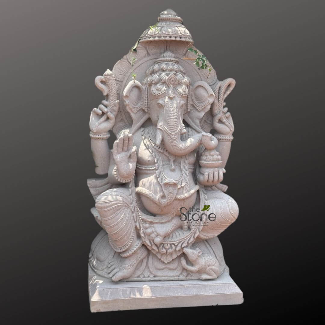 Big Ganesha Idol for Outdoor 4ft 3 majestic and intricately carved natural stone statue of Lord Ganesha in a luminous off-white or light grey hue is prominently displayed against a solid dark grey background, drawing full attention to its exquisite details. Ganesha is depicted seated comfortably on a rectangular plinth, with his right knee raised and his left leg folded, conveying a sense of serene presence and relaxed meditation. His iconic elephant head features large, fanned ears, a serene expression with half-closed eyes, and a prominent 'tilak' mark on his forehead. His trunk gracefully curves to his left ('Vamaturi Ganesha'), subtly touching a 'modak' (sweet dumpling) from the bowl held in his lower left hand. He is depicted with four arms, symbolizing his divine power. His upper right hand holds an 'Ankusha' (elephant goad), and his upper left hand grasps a 'Pasha' (noose), representing control and discipline. His lower right hand is raised in the 'Abhaya Mudra,' a gesture of fearlessness and blessing, while his lower left hand cradles the bowl of delicious modaks, symbolizing spiritual and material abundance. The sculpture is rich in decorative elements, showcasing a detailed tiered crown (mukuta), elaborate necklaces, armlets, bracelets, and anklets, all meticulously carved. A snake-like sacred thread ('Yagnopavitam' or 'Nagabandha') is draped across his portly belly, symbolizing wisdom and purity. His 'dhoti' (garment) is sculpted with flowing pleats and intricate patterns. At his feet, his 'vahana' (mount), a small rat or mouse (Mooshak), is sculpted, looking up attentively. Behind Ganesha, an ornate arch or 'Prabhavali' with delicate carvings frames his divine presence, accentuating his form. Small, verdant green vines subtly trail over the top of the crown and the Prabhavali, suggesting either the statue's integration with nature, its age, or a living, evolving reverence for the deity. This exquisite piece of Hindu art, likely crafted from high-quality marble or granite, exemplifies traditional Indian craftsmanship and makes a powerful statement as a spiritual focal point, garden statue, or decorative sculpture for a home, temple, or outdoor setting. It is a perfect representation for discussions on religious iconography, stone carving artistry, or spiritual home decor items.