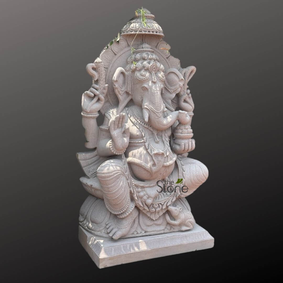 Big Ganesha Idol for Outdoor 4ft 2 majestic and intricately carved light gray stone statue of Lord Ganesha, the elephant-headed Hindu deity, is presented front and center against a plain, dark gray background. The entire sculpture exudes a sense of serene dignity and traditional craftsmanship. Ganesha is depicted seated in a dignified Lalitasana pose (royal ease), with his right leg folded and his left leg bent, resting on a sculpted double lotus pedestal (padmasana). His prominent elephant head features a gracefully curved trunk, large, detailed ears, and a single tusk (the right one appearing broken, a common iconographic detail). He wears an elaborate, layered crown adorned with intricate patterns, from which delicate green tendrils of a vine or plant gently emerge, suggesting an outdoor setting or natural growth. He possesses four arms, symbolizing his divine power and various attributes. His lower right hand is raised in the Abhaya Mudra, a gesture of blessing and reassurance, with finely sculpted fingers. His upper right hand holds an ankush (elephant goad), while his upper left hand grasps a mala (rosary beads) or pasha (noose). In his lower left hand, he cradles a small bowl of modak (sweet dumplings), a favorite treat. Ganesha's robust belly is adorned with a sacred thread (yagnopavit) sculpted in the form of a serpent, signifying his mastery over desires. He wears a variety of traditional jewelry, including necklaces, armlets, and bangles, all rendered with meticulous detail. The base of the sculpture is a beautifully carved double lotus, symbolizing purity and spiritual enlightenment. Near his left foot, his vahana (mount), a small, meticulously carved mouse (Mushika), sits attentively, looking up at Ganesha. Behind the deity, a richly detailed prabhavali (aura or archway) frames him, featuring traditional Indian floral and geometric motifs, topped with an ornate umbrella-like structure (chhatra). The uniform light gray tone of the stone highlights the exquisite texture and depth of the carving, showcasing the artistry involved in creating this traditional Hindu idol. This detailed Ganesha sculpture would be a powerful and auspicious addition to a garden, home temple, meditation space, or as a focal point in spiritual decor, symbolizing wisdom, prosperity, and the removal of obstacles.