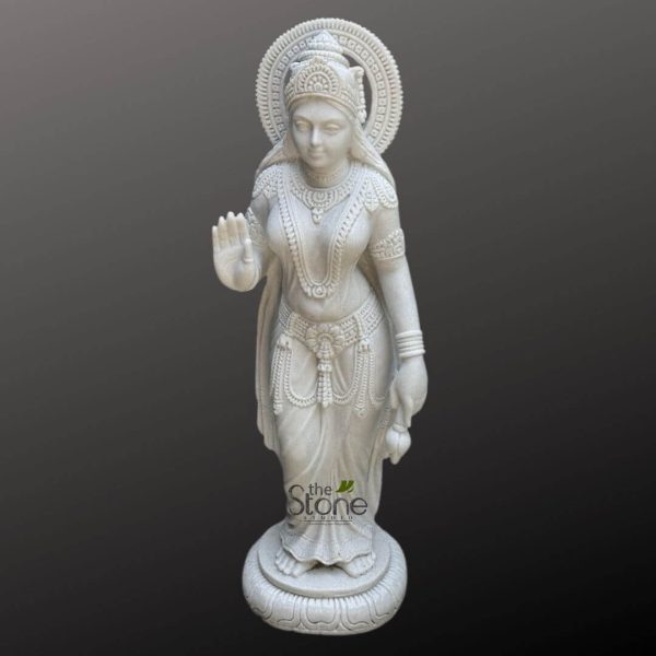 magnificent, full-length statue of a Hindu goddess, expertly crafted from pristine white marble, stands prominently against a plain, dark grey, almost black background. The goddess is depicted in a graceful, upright posture, emanating a sense of serenity and divine power, making her an ideal representation for devotional art or spiritual decor. Her head is adorned with an elaborate, multi-tiered crown, intricately carved with traditional motifs. A beautifully detailed circular halo, or Prabhamandal, frames her head, symbolizing her divine aura. Her face displays a peaceful and benevolent expression, with finely sculpted features including almond-shaped eyes, a delicate nose, and soft lips. Long, flowing hair is visible cascading from beneath her crown. She wears a rich array of traditional Indian jewelry, including prominent, multi-layered necklaces cascading down her chest, intricate earrings, elaborate armlets on her upper arms, and bangles on her wrists. Her slender form is draped in elegant, flowing robes, which are meticulously carved to show soft folds and textures, suggesting delicate fabric. A highly ornate waistband or hip-belt, adorned with decorative patterns and dangling elements, cinches her waist, further accentuating the exquisite craftsmanship of the marble sculpture. Her right hand is raised in the 'Abhaya Mudra,' a classical gesture of reassurance, blessing, and fearlessness, with the palm facing outwards and fingers extended upwards. Her left hand gently holds a delicate lotus bud or flower, which hangs naturally by her side, a common attribute of deities like Goddess Lakshmi or Saraswati. The goddess stands firmly on a beautifully carved lotus pedestal (padmasana), with intricate petal details visible around the circular base, further enhancing its spiritual significance. The immaculate polish and subtle grey veining inherent in the white marble highlight every minute detail of this hand-carved statue, showcasing superior artistic skill. The watermark "the Stone Studio" is subtly visible at the bottom center. This Hindu deity statue embodies grace, devotion, and masterful stone carving, suitable for a temple, meditation space, or a collector of sacred Indian art.