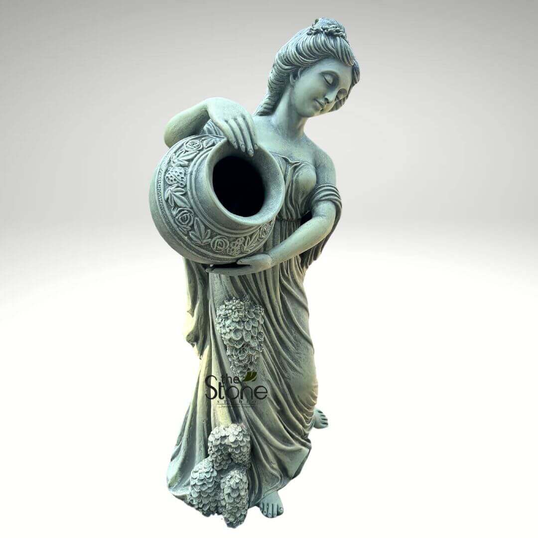 Water Fountain Lady Statue 5ft 4 full-length, classical-style garden statue crafted from a durable, weathered grey-green stone or cast concrete, depicting a graceful female figure. The statue stands upright, leaning slightly forward, with her head gently tilted downwards, eyes softly closed or cast in a serene, contemplative gaze, suggesting modesty or deep thought. Her hair is styled in an elegant, pulled-back fashion, possibly a bun, adorned with subtle floral or leaf-like embellishments that mimic a wreath or hair ornament. She is dressed in a flowing, floor-length gown characteristic of ancient Greek or Roman drapery, with soft folds and an off-the-shoulder neckline that reveals her bare shoulders and arms. Her bare feet are visible at the base, resting on an implied ground, adding to the classical aesthetic. With both hands, she carefully holds and tilts a large, rounded urn or pot forward, suggesting an act of pouring water or offering libations. The vessel is richly decorated with intricate, raised floral motifs, including detailed roses and swirling leaves, adding to its classical charm and craftsmanship. Notably, clusters of sculpted grapes or berries cascade elegantly down the front of her gown, particularly on her left side (viewer's right), emphasizing themes of nature, harvest, abundance, and perhaps a connection to deities like Persephone or a Bacchic celebration. The entire `stone sculpture` has a uniform, muted sage-green and grey tone, giving it an aged and classic appearance. This `garden feature` or `outdoor ornament` is presented against a plain, very light grey or white background, highlighting its form as a potential `focal point` for `landscape design`, `patio decor`, or `backyard aesthetics`. The detailed craftsmanship of this `classical statue` makes it a striking `outdoor art piece` ideal for enhancing any `home and garden` setting, particularly for those seeking a touch of timeless elegance or an `ancient-inspired outdoor sculpture`.