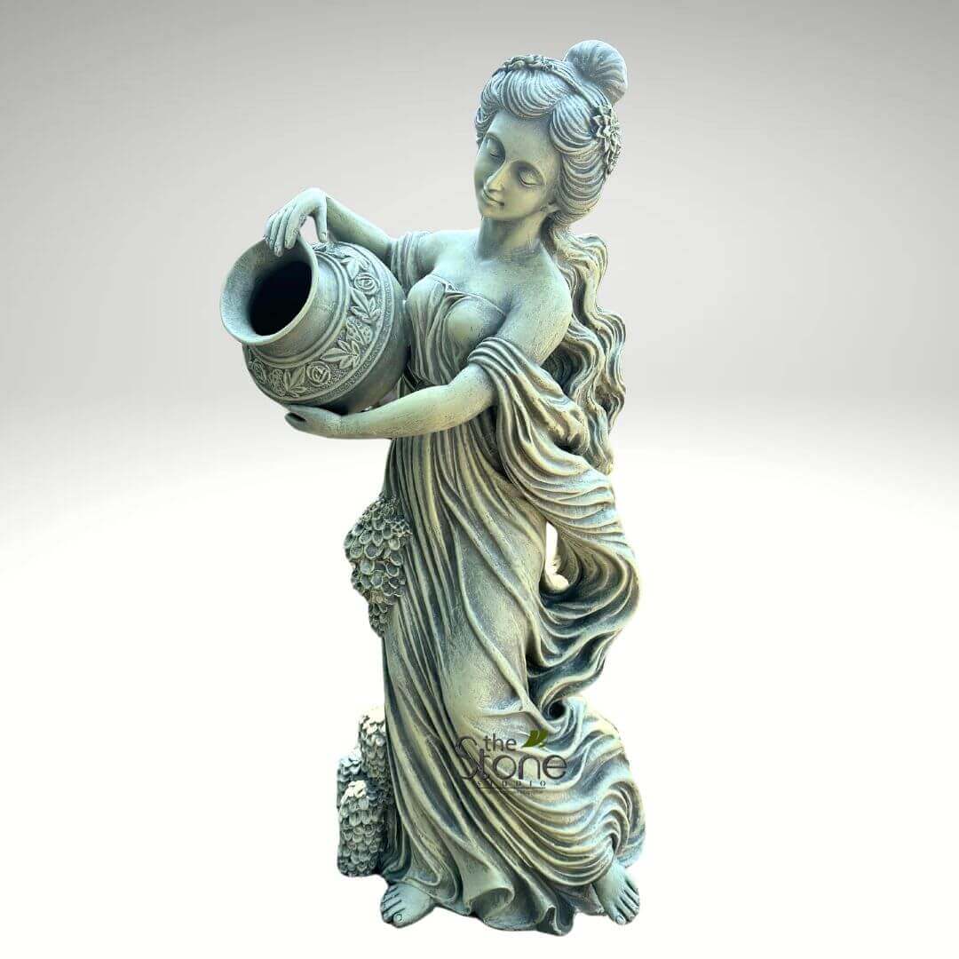 Water Fountain Lady Statue 5ft 1 magnificent, full-length classical garden statue depicting a graceful female figure, crafted from weathered stone or a similar durable material like concrete or cast cement. The statue stands tall, viewed from a slightly elevated angle against a plain, soft grey background that emphasizes its form and intricate details. The woman is elegantly dressed in flowing, neoclassical-style drapery that cascades around her body, creating soft folds and movement, especially around her legs and arms. Her posture is serene and poised, with her head gently tilted downwards and her eyes subtly closed or cast low, conveying a sense of calm contemplation or gentle effort, as if in the act of pouring. She holds a decorative, ornate amphora-like urn or jug with both hands, tipping it forward as if pouring water from its wide mouth. The urn itself is adorned with intricate relief carvings of floral or scroll patterns. Her hair is styled in an elaborate bun atop her head, secured with what appears to be a wreath or headband of small flowers, with long, wavy tresses falling gracefully over her shoulders and down her back. Intricate carvings of grapevines and clusters of grapes are visible along the lower portion of her gown and at the base of the statue, hinting at themes of abundance, nature, or possibly Dionysian symbolism. The entire piece exhibits a beautiful aged patina, a rich, muted green-grey color that suggests years of exposure to the elements, characteristic of antique garden ornaments or weathered outdoor sculptures. This exquisite stone art piece evokes timeless beauty and would be a striking focal point for landscaping projects, patio decor, or traditional garden designs, suitable as a water feature or a standalone decorative sculpture, emphasizing its craftsmanship and enduring appeal as a quality outdoor product.