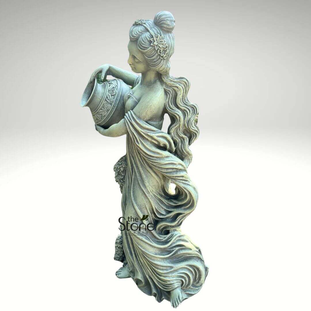 Water Fountain Lady Statue 5ft 2 full-length, highly detailed garden statue of a graceful female figure, crafted from what appears to be weathered cast stone or a similar durable resin, stands against a plain, light gray background. The sculpture boasts an antique finish, exhibiting subtle greenish-tan undertones that suggest age or the presence of moss, enhancing its classical aesthetic. The woman is depicted in a dynamic, poised stance, leaning gently forward as if in motion, with her head subtly bowed and gaze directed towards an ornate classical urn or pitcher she holds with both hands. Her long, wavy hair cascades down her back, while a portion is elegantly styled into an updo or bun, adorned with delicate floral or leafy details that interweave into her locks. She is clad in a flowing, draped gown, reminiscent of a Greco-Roman chiton, that gathers in soft, voluminous folds around her body and trails elegantly towards her bare feet, conveying a sense of graceful movement and classical artistry. The pitcher she holds features intricate raised relief patterns, possibly depicting botanical motifs, geometric designs, or classical figures, further enriching its ancient-inspired appearance. She gently tilts the vessel, implying the act of pouring water or making an offering. This exquisite piece, embodying the serene beauty of a mythological figure, a nymph, or a water bearer, is an ideal outdoor decor element. It would serve as a stunning focal point for a garden, patio, or courtyard, symbolizing abundance, tranquility, hospitality, or the life-giving essence of water. A watermark for "The Stone Studio" is visible on the lower left side of the statue, highlighting its origin and craftsmanship.
