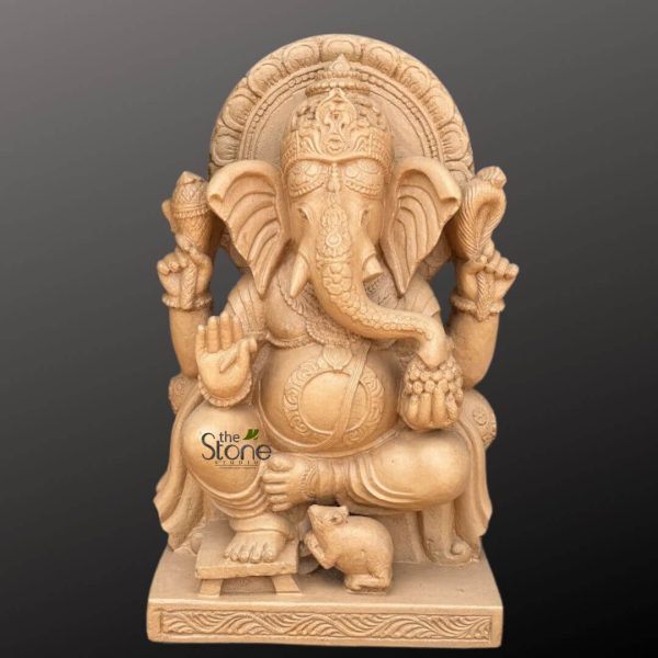 beautifully detailed, light beige or sandy-colored carved stone statue of the Hindu deity Lord Ganesha stands prominently against a simple, dark grey background. The entire sculpture exudes a sense of traditional Indian artistry and fine craftsmanship. Ganesha is depicted in a regal yet relaxed seated posture, with his left leg bent and his right foot resting comfortably on a small, ornate stool. He possesses his characteristic elephant head with large, fanning ears, intricately textured like elephant skin, and a gracefully curved trunk that bends towards his lower left hand. This hand holds a small cluster of round modakas (sweet dumplings), symbolizing blessings, prosperity, and the sweetness of spiritual attainment. Above the modakas, his upper left hand distinctly holds a coiled cobra with its hood raised, a powerful symbol of cosmic energy and protection. On his right side (viewer's left), his upper hand holds an ankusha (elephant goad), representing control over obstacles and the ability to guide devotees towards righteousness. His lower right hand is raised in the Abhaya Mudra, a universal gesture of fearlessness, reassurance, and blessing. Lord Ganesha wears an intricately carved mukuta (crown) on his head, adorned with delicate patterns, and various pieces of jewelry, including necklaces, armlets, and wristbands, all meticulously sculpted in the stone. His large, prominent belly, signifying abundance and the ability to consume all sorrows, is adorned with a decorative sash. Behind his head is a magnificent prabhamandala or aura, featuring ornate, radiating patterns that symbolize his divine energy and enlightenment. At his feet, nestled near his right foot, sits his faithful vahana (mount), a small, alert mouse, also carved with remarkable precision and detail. The entire handcrafted sculpture rests on a sturdy, multi-tiered rectangular pedestal, which is also adorned with subtle, repeating patterns. The stone has a smooth, polished finish, yet its natural texture and the impressive depth of the carvings are evident under the soft, even lighting, creating subtle shadows that enhance the three-dimensional quality of the piece. The warm, earthy tone of the stone makes this statue a perfect representation of peace, strength, and spiritual elegance. This image would be ideal for articles discussing Hindu art, spiritual home decor, Ganesha symbolism (such as the remover of obstacles, god of wisdom, and new beginnings), Vastu principles, or the beauty of handcrafted stone sculptures.