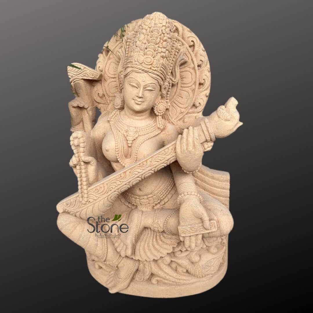 Saraswati Maa Statue 2.5ft 5 majestic and intricately carved stone sculpture, depicting the Hindu Goddess Saraswati, fashioned from light beige or sand-colored natural stone, possibly sandstone or marble, showcasing exquisite craftsmanship. The goddess is seated serenely in a traditional cross-legged meditative posture (Padmasana), emanating an aura of peace and wisdom. Her face bears a gentle, contemplative expression with soft, half-closed eyes and a subtle smile, conveying profound knowledge and tranquility. She wears an ornate, multi-tiered crown (mukut) richly adorned with delicate patterns and motifs, complemented by large, decorative ear ornaments. Saraswati is depicted with four arms, a common attribute of Hindu deities. Her two main hands delicately hold and play the Veena, a traditional Indian stringed instrument, which rests diagonally across her lap. The Veena itself is intricately carved with decorative scrollwork along its body. Her upper right hand cradles a Pustaka (sacred manuscript or book), symbolizing knowledge, learning, and literature, while her upper left hand holds a Japamala (rosary beads), signifying meditation, spirituality, and devotion. She is elaborately adorned with necklaces, armlets, bracelets, and a waist belt, all meticulously carved to suggest fine jewelry. Flowing drapery drapes gracefully around her lower body, with visible folds and textures, further enhancing the dynamic realism of the static stone. Behind her head, a prominent circular Prabhamandala (halo or aura) is exquisitely carved with radial lines and traditional Hindu patterns, emphasizing her divine presence and celestial nature. The entire sculpture rests on a simple, rounded base. The statue is presented against a plain, dark grey or black background, which dramatically highlights the light stone and the intricate details of the carving, drawing the viewer's complete attention to the artistry and the subject. A subtle watermark for "The Stone Studio" is visible on the lower left, indicating the origin or artisan behind this beautiful handcrafted idol. This piece represents traditional Indian iconography and artistry, embodying the essence of the goddess of knowledge, music, art, and wisdom, making it ideal for spiritual spaces, meditation rooms, home decor, or as an elegant centerpiece for cultural and religious appreciation.