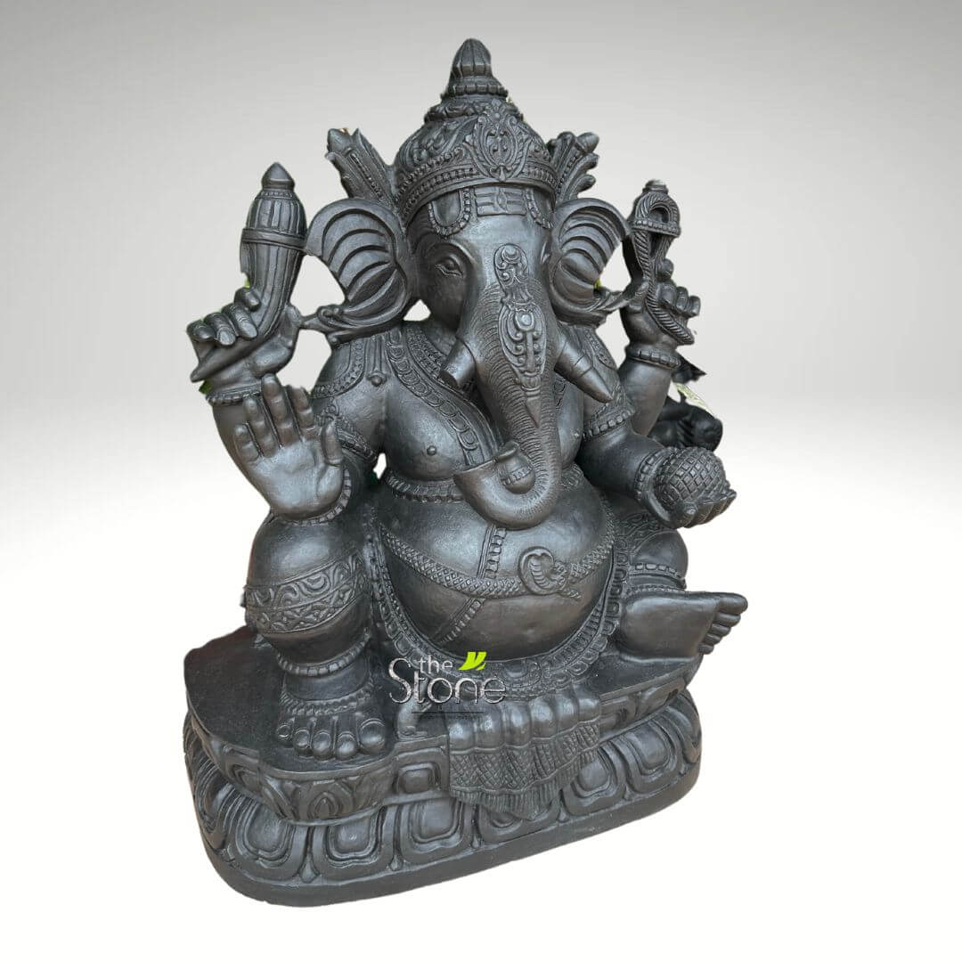 2.5 feet Stone Ganesha Statue 5 magnificent and intricately carved dark stone statue of Lord Ganesha is depicted, seated regally in a traditional meditative or royal posture (Padmasana) on a multi-tiered lotus pedestal. The elephant-headed Hindu deity is rendered in a deep charcoal grey or black, suggestive of polished basalt or a similar dense stone, with a rich, textural quality that highlights the detailed craftsmanship. Ganesha's portly belly, symbolizing prosperity and abundance, is prominent. His large, fanned ears are attentively open, and his gracefully curled trunk extends to the left, holding a sweet treat, likely a modak, which he is known to favor. He has four arms, each holding symbolic objects: his upper right hand clasps an ankusha (elephant goad), while his upper left holds a paasa (noose). His lower right hand is raised in the protective Abhaya Mudra, offering blessings and dispelling fear, with the palm facing forward. The lower left hand holds a small bowl or another modak, representing the rewards of spiritual endeavor. The statue is adorned with an elaborate, tiered crown, multiple necklaces, armlets, anklets, and a sacred thread (janeu) draped across his chest, all meticulously carved with traditional Indian patterns. His features, including the single visible tusk (as Ganesha is often depicted with one broken tusk), are rendered with profound artistry. The multi-layered lotus base provides a stable and aesthetically pleasing foundation, reflecting sacred symbolism. The entire sculpture is presented against a clean, light grey gradient background, which serves to emphasize the statue's form and intricate details without distraction. A subtle "The Stone" logo appears in green and white at the bottom center, likely indicating the artisan or material used. This detailed representation of Lord Ganesha, a remover of obstacles and bestower of wisdom and good fortune, is ideal for those seeking spiritual decor, a significant piece of Indian art, or a focal point for devotion in a home or garden setting.
