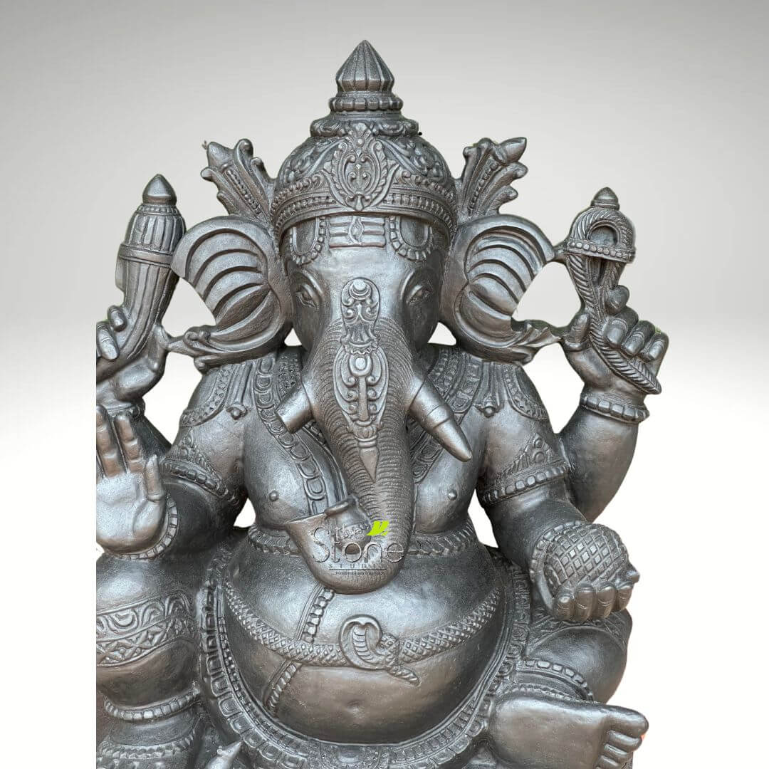 2.5 feet Stone Ganesha Statue 4 large, highly detailed, and exquisitely crafted statue of Lord Ganesha, the revered elephant-headed Hindu deity, stands prominently against a plain, light grey background. The sculpture boasts a dark metallic finish, possibly bronze or iron with an antique patina, giving it a rich, solemn, and aged appearance, with subtle lighter reflections highlighting its intricate textures. Lord Ganesha is depicted seated in a relaxed Lalitasana pose, with his right leg bent and drawn close to his body, and his left leg gently resting forward, conveying a sense of serene wisdom and approachable divinity. His iconic elephant head features large, fan-like ears adorned with delicate, patterned borders. His trunk curves gracefully towards his left, reaching down to gently touch a small modak (sweet dumpling) held in his lower left hand, symbolizing the rewards of spiritual endeavor and his love for sweets. Notably, Ganesha is depicted with one full tusk on his right side and a shorter, broken tusk on his left, a characteristic detail from Hindu mythology related to his role as a scribe. A majestic, multi-tiered crown, intricately carved with traditional Indian motifs including floral patterns and geometric designs, adorns his head, signifying his divine royalty. His serene eyes, almost almond-shaped, convey a profound sense of inner peace. The deity is traditionally depicted with four arms, each holding symbolic objects. His upper right hand gracefully holds a pasha (noose), representing the power to draw devotees closer to the divine and control worldly attachments. His upper left hand grasps an ankusha (goad), symbolizing the ability to guide and clear obstacles. His lower right hand is raised in the abhaya mudra, a gesture of reassurance, protection, and fearlessness for his devotees. As mentioned, his lower left hand holds the modak towards his trunk. Ganesha's robust, pot-bellied form, a symbol of prosperity, generosity, and the ability to consume sorrows, is richly adorned with elaborate traditional Indian jewelry. These include multiple necklaces with beaded and coiled snake designs, ornate armlets, anklets, and a ceremonial waistband. The dhoti-like drapery around his lower body is also intricately textured with patterns. The entire statue, from the delicate patterns on his crown and jewelry to the subtle contours of his trunk and body, demonstrates exceptional craftsmanship, reflecting the deep cultural and spiritual significance of this Hindu god of wisdom, new beginnings, and the remover of obstacles. This evocative image is perfect for articles discussing Hindu mythology, spiritual practices, traditional Indian art, cultural heritage, home decor, or the symbolic representation of prosperity and good fortune.