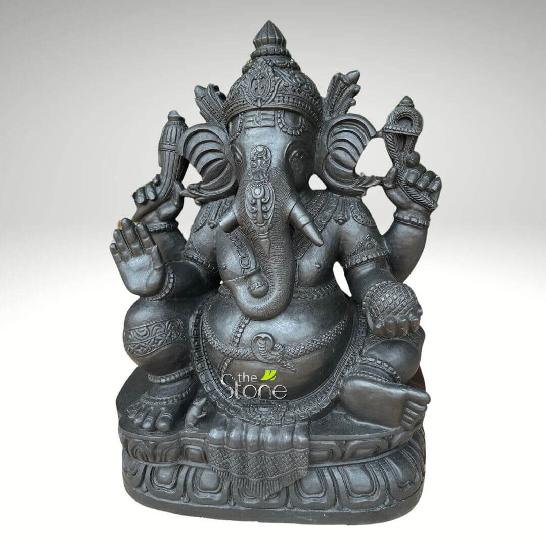 2.5 feet Stone Ganesha Statue 3 majestic and intricately detailed dark charcoal grey stone statue of Lord Ganesha is presented front and center against a neutral, softly lit light grey background, highlighting its exquisite craftsmanship. The revered Hindu deity, known as the remover of obstacles, is depicted seated in a relaxed, regal posture, often referred to as Lalitasana, with one leg folded and the other slightly lowered, symbolizing ease and grace. Ganesha's iconic elephant head is prominent, featuring large, fanned ears and a gracefully curved trunk that touches or holds a small item, possibly a sweet (modak or laddu), or simply an elegant curl. He wears an elaborate, tiered crown (mukuta) adorned with complex patterns, and his forehead bears sacred marks. One of his tusks appears broken, a traditional characteristic linked to tales of wisdom and sacrifice. The statue showcases Ganesha's four arms, each holding symbolic attributes. His lower right hand is raised in the "Abhaya Mudra," a gesture of blessing, reassurance, and fearlessness. The upper right hand holds an "ankusha" (goad), while the upper left hand grasps a "pasha" (noose), representing control over desires and attachments. His lower left hand rests gently on his knee, possibly holding a small offering or rosary beads. Ganesha's generous, pot-bellied form, symbolizing prosperity and bounty, is adorned with numerous finely carved necklaces, armlets, bracelets, and anklets, all featuring intricate designs. A sacred thread, resembling a coiled serpent (Yajnopavitam), drapes elegantly across his chest and torso. His draped garment (dhoti) is also richly textured with patterns. He sits upon a beautifully sculpted, multi-layered lotus pedestal (Padmasana-style base), which itself is intricately patterned with stylized petals and geometric motifs, elevating the deity. The overall finish of the statue is a deep, uniform dark grey, suggesting a high-quality, polished stone such as granite or basalt, giving it an antique yet refined appearance. A subtle watermark "the Stone" is visible on the lower front, reinforcing the material and potential artisan origin. This stone carving embodies the spiritual essence of Ganesha, the god of wisdom, prosperity, and new beginnings, making it an ideal representation for spiritual spaces, home decor, or as a focal point for devotion. The meticulous detail and substantial material reflect enduring artisan craftsmanship and cultural significance, connecting naturally to articles discussing Hindu deities, traditional Indian art, stone sculptures, or high-quality spiritual artifacts.