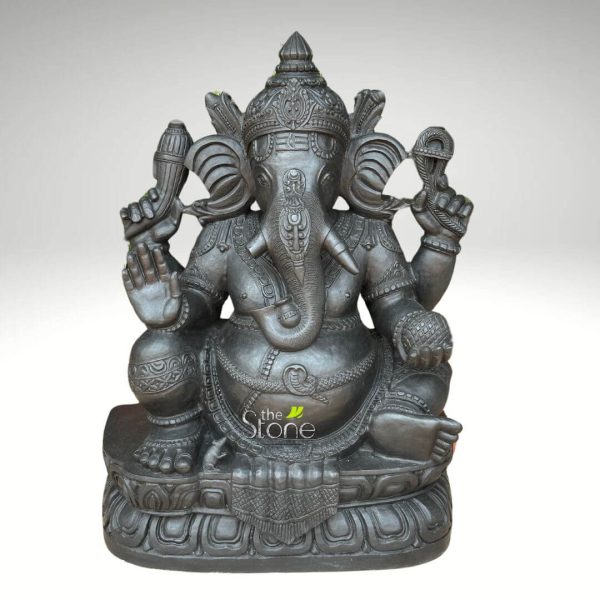 magnificent and intricately carved, dark charcoal gray stone sculpture of the Hindu deity Lord Ganesha, depicted in a regal, seated posture against a clean, light gray background. The substantial statue showcases exquisite craftsmanship, likely in granite or black marble, with a rich, matte finish that highlights every detail of the traditional Indian artistry. Ganesha, the elephant-headed god revered as the remover of obstacles (Vighnaharta) and bestower of wisdom, prosperity, and success (Siddhivinayak), is shown with his iconic four arms. His prominent elephant head features large, fan-like ears and a trunk elegantly curled towards his right, with a single, unbroken tusk (ekadanta) symbolizing intellectual sacrifice and knowledge. He wears a majestic, multi-tiered crown (mukuta) adorned with elaborate carvings and patterns, along with finely detailed necklaces, armlets, and bracelets. His upper right hand (viewer's left) holds his broken tusk, while his lower right hand is raised in the Abhaya Mudra, a universal gesture of fearlessness, protection, and blessing. In his upper left hand (viewer's right), he holds an ankusha (elephant goad), representing divine guidance, control over worldly desires, and the path to righteousness. His lower left hand gently cradles a Modak, his favorite sweet, symbolizing rewards, spiritual liberation, and the sweetness of a realized soul. The deity's pot-bellied form, a symbol of abundance, generosity, and the cosmos, is adorned with a sacred thread (janeu) draped across his chest and a finely sculpted waistband. He sits comfortably in Lalitasana (royal ease pose) on a meticulously sculpted double-lotus pedestal (padmasana), which features continuous patterns of lotus petals and other traditional Hindu decorative motifs, signifying purity and spiritual enlightenment. The subtle "the Stone" watermark reinforces the material and suggests its relevance for articles on spiritual home decor, traditional Indian stone sculpture, or the symbolism and cultural significance of Hindu deities. This detailed piece serves as a powerful representation of devotion and artistic heritage.