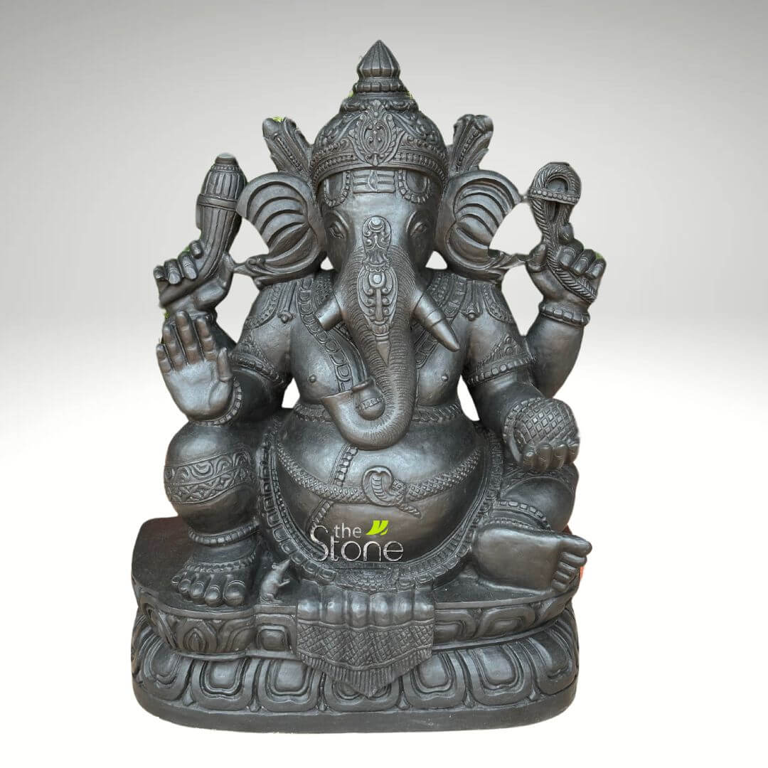 2.5 feet Stone Ganesha Statue 1 magnificent and intricately carved, dark charcoal gray stone sculpture of the Hindu deity Lord Ganesha, depicted in a regal, seated posture against a clean, light gray background. The substantial statue showcases exquisite craftsmanship, likely in granite or black marble, with a rich, matte finish that highlights every detail of the traditional Indian artistry. Ganesha, the elephant-headed god revered as the remover of obstacles (Vighnaharta) and bestower of wisdom, prosperity, and success (Siddhivinayak), is shown with his iconic four arms. His prominent elephant head features large, fan-like ears and a trunk elegantly curled towards his right, with a single, unbroken tusk (ekadanta) symbolizing intellectual sacrifice and knowledge. He wears a majestic, multi-tiered crown (mukuta) adorned with elaborate carvings and patterns, along with finely detailed necklaces, armlets, and bracelets. His upper right hand (viewer's left) holds his broken tusk, while his lower right hand is raised in the Abhaya Mudra, a universal gesture of fearlessness, protection, and blessing. In his upper left hand (viewer's right), he holds an ankusha (elephant goad), representing divine guidance, control over worldly desires, and the path to righteousness. His lower left hand gently cradles a Modak, his favorite sweet, symbolizing rewards, spiritual liberation, and the sweetness of a realized soul. The deity's pot-bellied form, a symbol of abundance, generosity, and the cosmos, is adorned with a sacred thread (janeu) draped across his chest and a finely sculpted waistband. He sits comfortably in Lalitasana (royal ease pose) on a meticulously sculpted double-lotus pedestal (padmasana), which features continuous patterns of lotus petals and other traditional Hindu decorative motifs, signifying purity and spiritual enlightenment. The subtle "the Stone" watermark reinforces the material and suggests its relevance for articles on spiritual home decor, traditional Indian stone sculpture, or the symbolism and cultural significance of Hindu deities. This detailed piece serves as a powerful representation of devotion and artistic heritage.