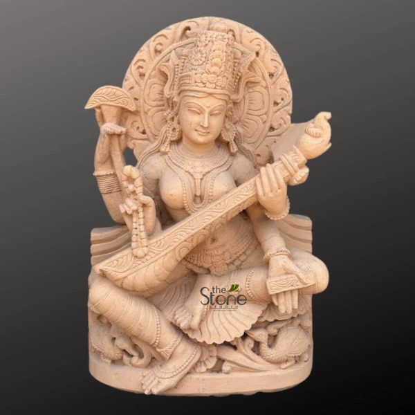 stunning, intricately carved sculpture of the Hindu Goddess Saraswati, crafted from a light-beige or sandstone-colored natural stone. The deity is depicted seated gracefully in a traditional pose, embodying serenity and wisdom. She possesses four arms, characteristic of many Hindu gods and goddesses. Her primary two hands delicately hold a traditional Indian stringed instrument known as a veena, with her fingers positioned as if playing a melody. In her upper right hand, she holds a string of prayer beads (mala), symbolizing meditation and spirituality. Her upper left hand holds a sacred palm-leaf manuscript or scroll, representing knowledge, scriptures, and learning. Saraswati is adorned with an elaborate crown (mukuta) featuring intricate floral and geometric patterns, and fine jewelry including necklaces, earrings, armlets, bracelets, and a waistband, all meticulously carved into the stone. Her facial expression is calm and benevolent, with softly sculpted features and downcast eyes. Behind her head, an ornate, circular prabhamandala (halo or aura) is visible, deeply carved with traditional motifs that enhance her divine presence. The goddess is seated on a sturdy, multi-layered pedestal, which features detailed carvings along its base. These carvings include depictions of swans (hamsa), Saraswati's sacred vehicle, nestled amongst flowing water or foliage patterns, symbolizing purity and discernment. The entire sculpture exhibits exceptional traditional Indian craftsmanship, with smooth, polished surfaces juxtaposed against highly detailed, textured adornments and drapery. The statue is presented against a plain, dark grey background, which effectively highlights the natural texture and warm tone of the stone, emphasizing its exquisite artistry and the spiritual significance of the Hindu deity. The watermark "the Stone Studio" is subtly visible on the lower right, indicating the source of this fine art piece. This sculpture beautifully represents the attributes of music, arts, knowledge, and wisdom that Goddess Saraswati embodies.