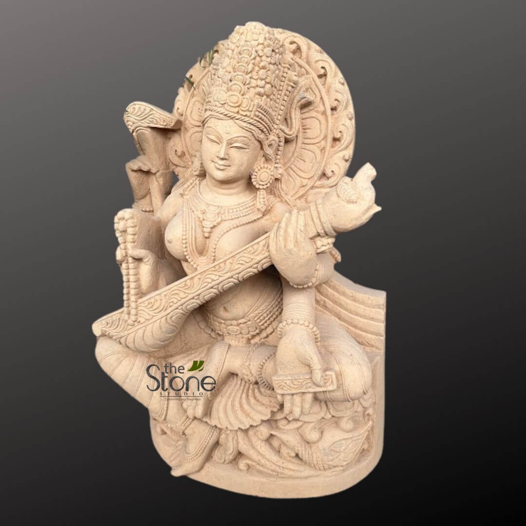 Saraswati Maa Statue 2.5ft 4 stunning, highly detailed cream-colored carved stone sculpture depicts Goddess Saraswati, the Hindu deity of knowledge, music, arts, wisdom, and learning. The multi-armed goddess is seated gracefully in a regal posture, centrally positioned against a plain dark gray background that accentuates the intricate craftsmanship. Her serene, slightly smiling face is adorned with delicate features, including soft lips and almond-shaped eyes, radiating tranquility. She wears an elaborate, tall crown (mukut) intricately carved with layers of decorative motifs, possibly resembling lotus petals or a sunburst, along with ornate earrings, multiple necklaces, armlets, and bracelets, all rendered with exquisite precision. Her upper body is adorned with garlands, and her lower body is draped in a flowing garment with finely sculpted pleats and decorative borders that cascade over her legs. Saraswati is depicted playing a veena, a long-necked string instrument. Her lower two hands are delicately positioned on the instrument, one plucking the strings and the other holding the fretboard, conveying a sense of harmonious movement. Her upper left hand gracefully holds a strand of mala (rosary beads), symbolizing concentration and meditation. Her upper right hand is poised in a delicate gesture near the top of the veena, perhaps ready to hold a pustaka (manuscript), which is often associated with her. Behind her head, a circular prabhavali (aureole or halo) is richly carved with swirling, almost flame-like or floral motifs, emphasizing her divine presence and aura. She rests upon a sculpted, multi-tiered lotus-like pedestal, which is also adorned with intricate patterns, providing a stable and sacred base. The entire sculpture showcases exceptional artistry, with every element, from the individual beads of the mala to the strings of the veena and the delicate patterns on her attire and the base, meticulously carved. The light, natural stone material highlights the fine textures and contours, giving the piece a timeless, elegant, and spiritual aesthetic. This exquisite piece of traditional Indian stone art beautifully captures the essence of divine wisdom and artistic expression. A subtle "The Stone Studio" watermark is visible on the lower left side of the sculpture.
