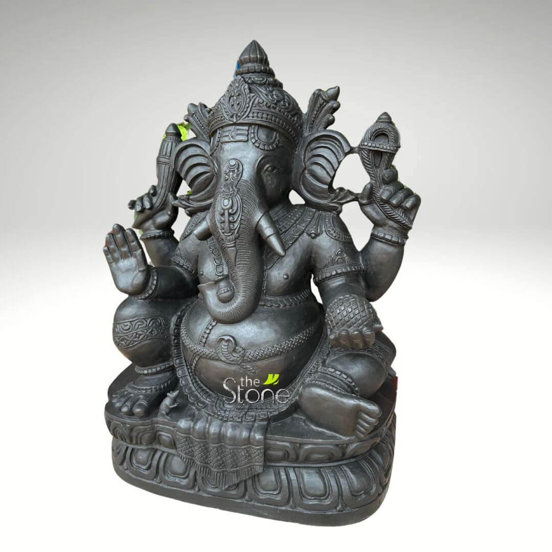 2.5 feet Stone Ganesha Statue 2 magnificent, highly detailed sculpture of Lord Ganesha, the revered Hindu deity with an elephant head, is prominently displayed against a clean, softly diffused light grey to white background. The statue is crafted from what appears to be dark charcoal grey stone or a similar durable, weighty material, possessing a matte finish that highlights its intricate carvings. Lord Ganesha is depicted seated in a serene and tranquil posture, often associated with meditation and blessing. He possesses four arms, each holding symbolic items: his front right hand is raised in the Abhaya Mudra, a gesture of fearlessness and bestowing blessings, with the palm facing forward. His front left hand gently holds a modak, a sweet dumpling, signifying the sweetness of spiritual realization and rewards. In his upper right hand, he grasps an ankusha (elephant goad), representing the ability to guide and remove obstacles, while his upper left hand holds a pasha (noose), symbolizing the restraint of worldly attachments. The deity's elephant head features large, fanning ears, small, focused eyes, and a gracefully curled trunk that turns towards his left. His iconic single tusk is visible, a symbol of sacrifice and wisdom. He is adorned with an elaborate crown (kiritamukuta) and a profusion of finely carved traditional jewelry, including necklaces, armbands, wristbands, and a detailed waistband that gracefully accents his portly belly, a symbol of prosperity and abundance. The sculpture rests upon a beautifully sculpted lotus pedestal (Padma Peetha), rich with petaled details, which in turn sits on a sturdy, layered rectangular base. The entire piece showcases exceptional craftsmanship, with every fold of drapery and ornamental pattern meticulously rendered. A subtle "The Stone" watermark is visible on the lower front portion of the statue, likely indicating the artisan or vendor. This striking piece embodies spiritual significance, traditional Indian artistry, and cultural heritage, making it an ideal subject for articles on Hindu mythology, sacred sculptures, home décor featuring religious idols, or the craftsmanship of stone carving.