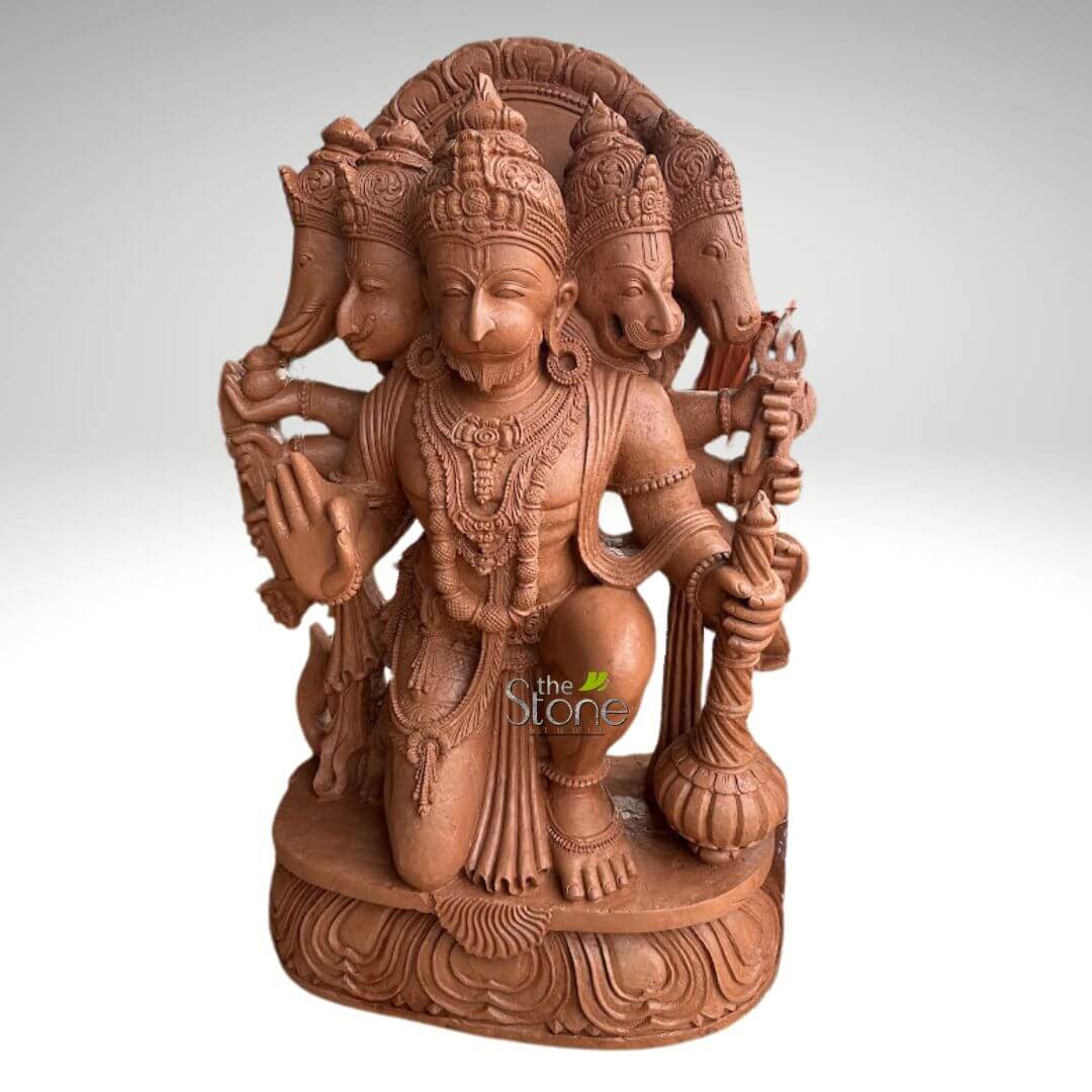 4 Feet Panchamukhi Hanuman Statue 5 detailed, reddish-brown stone sculpture depicts the multi-headed and multi-armed Hindu deity, Panchmukhi Hanuman, kneeling on an ornate lotus pedestal. The central and largest head is that of Hanuman, featuring a serene expression, closed eyes suggesting meditation or devotion, a prominent moustache, and an elaborate crown (mukuta). Surrounding the main Hanuman face are four other divine heads, representing different manifestations: to the viewer's immediate left is the elephant head of Lord Ganesha, and further left, the eagle-like head of Garuda; to the viewer's immediate right is the fierce lion face of Lord Narasimha, and further right, the boar head of Lord Varaha. Hanuman is portrayed in a powerful yet reverent kneeling stance, with his right knee on the ground and his left foot firmly planted on the base. His numerous arms hold various divine attributes: in his prominent front right hand, he grasps a heavy mace (gada), symbolizing strength and protection, while his front left hand is raised in the Abhaya Mudra, a gesture of fearlessness and blessing. Other hands are seen holding a trident (trishul) and possibly a noose or rope. The entire sculpture is intricately carved from a matte, terracotta-colored stone, showcasing exquisite traditional Indian craftsmanship. Every detail, from the ornate crowns, necklaces (mala), armbands, and anklets adorning the deity to the flowing dhoti and the finely chiseled expressions of each face, is rendered with precision. The deep, earthy hue of the stone accentuates the ancient and spiritual essence of the artwork, highlighting its connection to Hindu mythology and devotional practices. The sculpture is set against a plain, light grey background, allowing its intricate details and spiritual significance to be the sole focus, making it a prominent piece for worship or a significant representation of divine power and protection.
