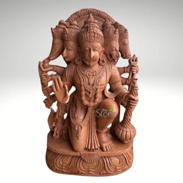 meticulously carved reddish-brown stone sculpture depicts the Panchmukhi Hanuman, a multi-faced form of the Hindu deity Hanuman, kneeling powerfully on an ornate lotus pedestal. The central figure is Hanuman, rendered with a strong, muscular physique, wearing a detailed crown and an elaborate collection of traditional Indian jewelry, including multiple necklaces, armbands, and a sacred thread (yajnopavita). His own monkey-like face, with a serene yet determined expression and a neatly carved beard, is at the center. Flanking him are two other prominent divine heads: Lord Ganesha (the elephant-headed deity) on his left, and Lord Narasimha (the fierce lion-headed avatar of Vishnu, depicted with an open mouth and fangs) on his right. Behind Ganesha's head, the eagle-headed Garuda is visible, while behind Narasimha, the horse-headed Hayagriva completes the five faces of this powerful Panchmukhi manifestation. Hanuman is shown with ten arms, each adorned with intricate bracelets and armbands. His front left hand is raised in the gesture of blessing (Abhaya Mudra), signifying fearlessness and protection. In his other hands, he firmly grasps various divine weapons and symbols: most notably, a prominent mace (gada) and a trident (Trishul) are visible on his right side, along with a bow and arrow held on his left. The meticulous carving extends to the details of his dhoti (waist cloth) and the flowing sash behind him. The entire divine assembly rests on a multi-layered, intricately carved lotus pedestal, symbolic of purity and spiritual awakening. The reddish-brown material, likely sandstone or a terracotta-like stone, showcases exceptional craftsmanship, highlighting the texture and depth of the traditional Indian stone carving. The sculpture is set against a plain, light grey background, allowing its intricate details and spiritual presence to stand out, making it an ideal representation of Hindu devotion and artistry suitable for spiritual spaces, home altars, or as a magnificent example of cultural heritage.