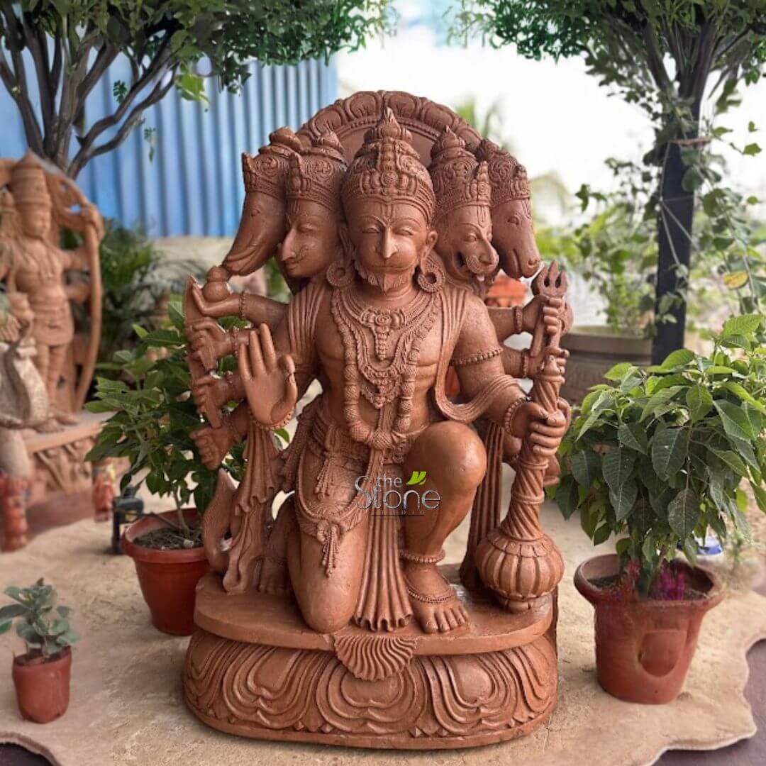 4 Feet Panchamukhi Hanuman Statue 2 large, intricately carved reddish-brown stone sculpture depicting Panchamukhi Hanuman, the five-faced manifestation of Lord Hanuman, is prominently displayed outdoors. The central figure of Hanuman is shown in a dynamic, kneeling posture on his right knee, with his left foot firmly planted on an ornate, multi-tiered lotus petal base. He is powerfully built, with a muscular torso and adorned with multiple arms. His primary front right hand firmly grips a formidable gada (mace) with a detailed twisted handle, while his primary front left hand is raised in the Abhaya Mudra, signifying fearlessness, blessing, and protection. Other hands hold a bow with an arrow, and a trident (trishul), symbolizing his divine strength and various attributes. Above Hanuman's central, crowned monkey face, which features a detailed beard and expressive eyes, are four additional, intricately sculpted heads, completing the Panchamukhi (five-faced) form. These heads, from left to right, include: a benevolent elephant head (representing Ganesha or Gaja Mukha), a fierce, human-like head with sharp features (possibly Narasimha or Hayagriva), a majestic horse head (representing Hayagriva), and a powerful boar's head (representing Varaha). Each of these divine faces is adorned with its own crown or elaborate headgear, and behind them, an intricate, fan-shaped arch or halo adds to the sculpture's grandeur. The entire statue is meticulously detailed with traditional Hindu iconography, including layered necklaces, prominent earrings, armbands, wristbands, and anklets. His dhoti is sculpted with realistic folds and patterns. The reddish-brown hue of the sculpture suggests a natural stone material like terracotta or sandstone, highlighting the fine craftsmanship and texture. The statue is positioned in a serene outdoor setting, surrounded by lush green foliage. Numerous potted plants of various sizes, with vibrant green leaves, are arranged around its base, enhancing the natural ambiance. In the background, taller trees provide a canopy of green, and to the left, a glimpse of a faint blue corrugated structure suggests a garden, a traditional courtyard, or an outdoor studio display area for such spiritual artworks. This visually striking piece embodies themes of divine power, protection, spiritual devotion, and the rich tradition of Indian mythological sculpture, making it a compelling image for articles on Hindu deities, stone carving, religious art, or sacred symbolism.