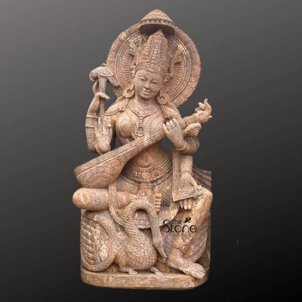 magnificent, intricately carved light brown sandstone sculpture depicts the revered Hindu Goddess Saraswati, the deity of knowledge, music, arts, wisdom, and learning. She is seated gracefully in a meditative posture, adorned with an elaborate, finely detailed crown and numerous pieces of traditional Indian jewelry, including necklaces, armlets, bangles, and large earrings. Her serene face, with finely sculpted features and a gentle, welcoming smile, radiates peace and wisdom. The goddess is depicted with four arms, a common attribute for Hindu deities, symbolizing her multifaceted abilities. Her two primary hands delicately play a beautifully ornamented veena, a traditional South Indian string instrument, whose long neck and resonant body are intricately carved with decorative patterns. Her upper right hand holds a mala (rosary beads), representing spiritual discipline and meditation, while her upper left hand grasps a pustaka (sacred manuscript or book), symbolizing knowledge and scriptures. Behind her head, a large, circular prabhamandala (aura or halo) with concentric decorative patterns rises, topped by a smaller conical spire, emphasizing her divine presence. At the base of the sculpture, to her left, her celestial vehicle (vahana), a majestically carved hamsa (swan), is depicted with its neck elegantly curved, looking up towards the goddess. The swan symbolizes purity, discernment, and the ability to distinguish between good and evil. The entire stone sculpture showcases exceptional craftsmanship, with every fold of drapery, every jewel, and every feather of the swan rendered with remarkable precision and depth. The natural texture and warm, light brown hues of the sandstone material give the piece an earthy yet ethereal quality. This exquisite piece, possibly crafted by "the Stone studio" as indicated by the watermark, stands against a plain, dark grey background, allowing its intricate details and spiritual significance to stand out prominently. It serves as a powerful representation of traditional Indian art, spiritual devotion, and cultural heritage, perfect for those seeking a handcrafted deity idol, a spiritual art piece, or an emblem of wisdom and creativity.