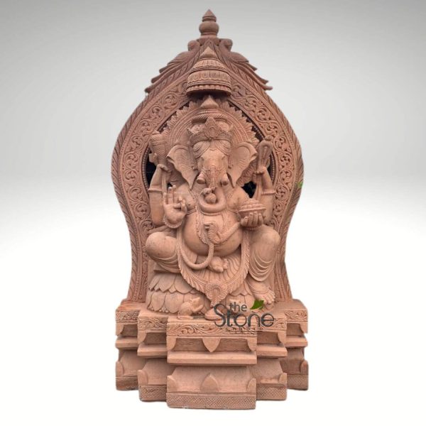 meticulously carved, reddish-brown sandstone statue of Lord Ganesha, presented as a full-view product image against a plain, light gray studio background. Lord Ganesha is depicted seated in a majestic, benevolent cross-legged posture (possibly Padmasana or Sukhasana) on a multi-tiered lotus pedestal (Padma Peetha). He features his iconic elephant head with large, fanned ears, small, serene eyes, and a gracefully curved trunk turned towards his left, gently touching a modak in his lower left hand. He has a single, broken tusk, symbolizing wisdom and sacrifice, and a prominent, round belly, indicative of prosperity and abundance. Ganesha is adorned with an elaborate, tiered crown (mukuta), multiple necklaces, armlets, bracelets, and anklets. A sacred thread (Yagnopavitam) is creatively fashioned as a coiled snake across his chest. He possesses four arms: his upper right hand holds an ankusha (elephant goad), the upper left a pasha (noose), his lower left hand is raised in the Abhaya mudra, offering blessings and protection, and his lower right hand holds a small bowl filled with his favorite sweet dumplings, modaks. Behind Ganesha, a magnificent and intricately carved archway, known as a Prabhavali, rises upwards, framing the deity. This ornate backdrop is adorned with concentric layers of finely detailed floral motifs, swirling vine patterns, and geometric scrollwork, culminating in a tiered, spire-like finial at the very top. A circular, halo-like design with sunburst patterns frames Ganesha's head, highlighting his divine aura. At Ganesha's feet, his faithful vahana, the mouse (Mushika), is depicted seated, looking up towards him, possibly also holding a tiny modak. The entire sculpture rests upon a sturdy, multi-tiered, rectangular pedestal that complements the main figure with simple yet elegant decorative moldings. The sculpture's warm, earthy reddish-brown hue and visible texture suggest it is crafted from high-quality sandstone, showcasing exceptional traditional Indian stone carving artistry and intricate detail across all elements, from the deity's features to the decorative patterns of the Prabhavali. A subtle "the Stone Studio" watermark is visible on the lower front of the pedestal. This detailed Ganesha idol is an embodiment of wisdom, prosperity, and the remover of obstacles, ideal for spiritual worship, home decor, or as a significant piece of cultural art.