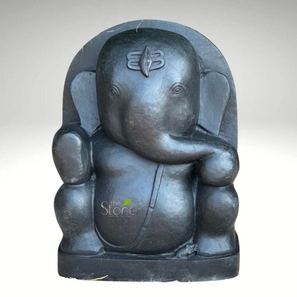 majestic and finely crafted dark stone statue of Lord Ganesha, the revered Hindu deity, stands centrally against a clean, plain light grey background. The sculpture, likely carved from a deep grey or black stone like basalt or granite, possesses a smooth, matte finish that highlights its rounded, robust forms. Lord Ganesha is depicted in a serene, seated posture, conveying an aura of peace and contemplation. His iconic elephant head features large, gracefully curved ears and a prominent trunk that curls gently towards his left, resting near his rotund belly, a common representation of his prosperity and wisdom. His eyes are subtly carved, appearing half-closed, adding to his meditative expression. A distinct and detailed 'tripundra' mark, a Shiva tilak symbolizing spiritual wisdom and devotion, is prominently carved on his forehead. He is shown with two visible arms; his right arm is bent at the elbow, with the hand resting near his chest, while his left hand rests comfortably on his left knee. A slender, simple sash or sacred thread (upavita) elegantly drapes across his left shoulder and down his torso, emphasizing his divine status. The deity is seated upon a sturdy, integrated rectangular base, with a solid, arched backrest rising behind him, framing his upper body and adding to the sculpture's monolithic feel. The overall design is traditional yet appears to have a simplified, elegant aesthetic, emphasizing Ganesha's benevolent form as the Remover of Obstacles and the Lord of Beginnings. The statue's solid craftsmanship and detailed yet understated features make it an ideal representation of Hindu spiritual art, perfect for articles discussing stone carving techniques, religious iconography, traditional Indian handicrafts, or as a focal point for spiritual home and garden decor. A subtle watermark, "the Stone," is visible on the lower left, indicating the material or maker.
