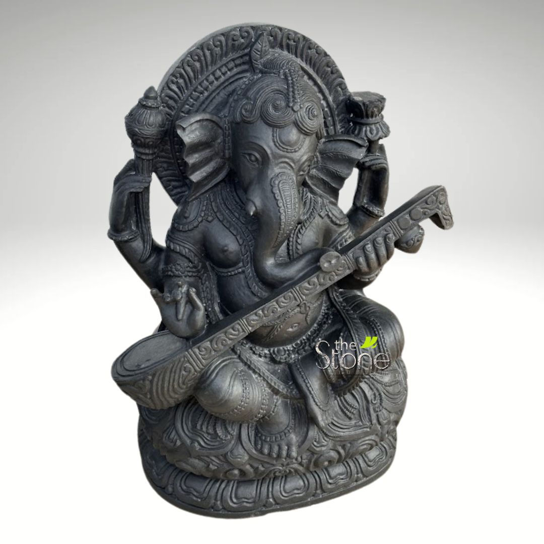 Veena Ganesha Statue 2ft 5 detailed and intricately carved dark grey stone statue depicts the Hindu deity Ganesha, the elephant-headed god of wisdom, prosperity, and remover of obstacles. Ganesha is seated in a relaxed and graceful posture, with one leg drawn up and the other slightly extended, appearing to rest on a beautifully sculpted lotus pedestal. His iconic elephant head features large, fan-like ears, small eyes, and a trunk curled gently to his left, almost touching his pot belly. The four-armed deity is richly adorned with traditional Indian jewelry, including layered necklaces, elaborate armlets, wristlets, and anklets, and a decorative waist belt. A multi-tiered crown sits atop his head. His upper left hand holds a stylized weapon or staff, possibly a gada, while his upper right hand holds what appears to be a modak (a sweet dumpling) or a lotus bud, symbolizing offerings or blessings. His lower left hand is positioned in a serene mudra (blessing gesture) resting on his knee, while his lower right hand delicately plucks the strings of a long, elaborately carved musical instrument resembling a veena, which rests across his lap. The veena itself is adorned with intricate patterns, emphasizing Ganesha's role as the divine musician and patron of arts. Behind Ganesha's head, an ornate, arch-shaped prabhavali (aura or halo) features detailed radial and floral carvings, enhancing the divine presence of the sculpture. The entire statue is rendered in a dark, matte finish, characteristic of high-quality stone such as granite or basalt, giving it a heavy, earthy, and ancient aesthetic. The texture appears slightly rough yet refined, highlighting the artisan's skill in stone carving. The statue is well-lit against a clean, light grey to white gradient background, which emphasizes its three-dimensional form and detailed craftsmanship. The watermark "the Stone" subtly reinforces the material and potential source, suggesting the article might focus on stone artistry, religious sculptures, Indian cultural decor, or handcrafted deities. This piece embodies spiritual artistry, musical harmony, and traditional Indian craftsmanship.