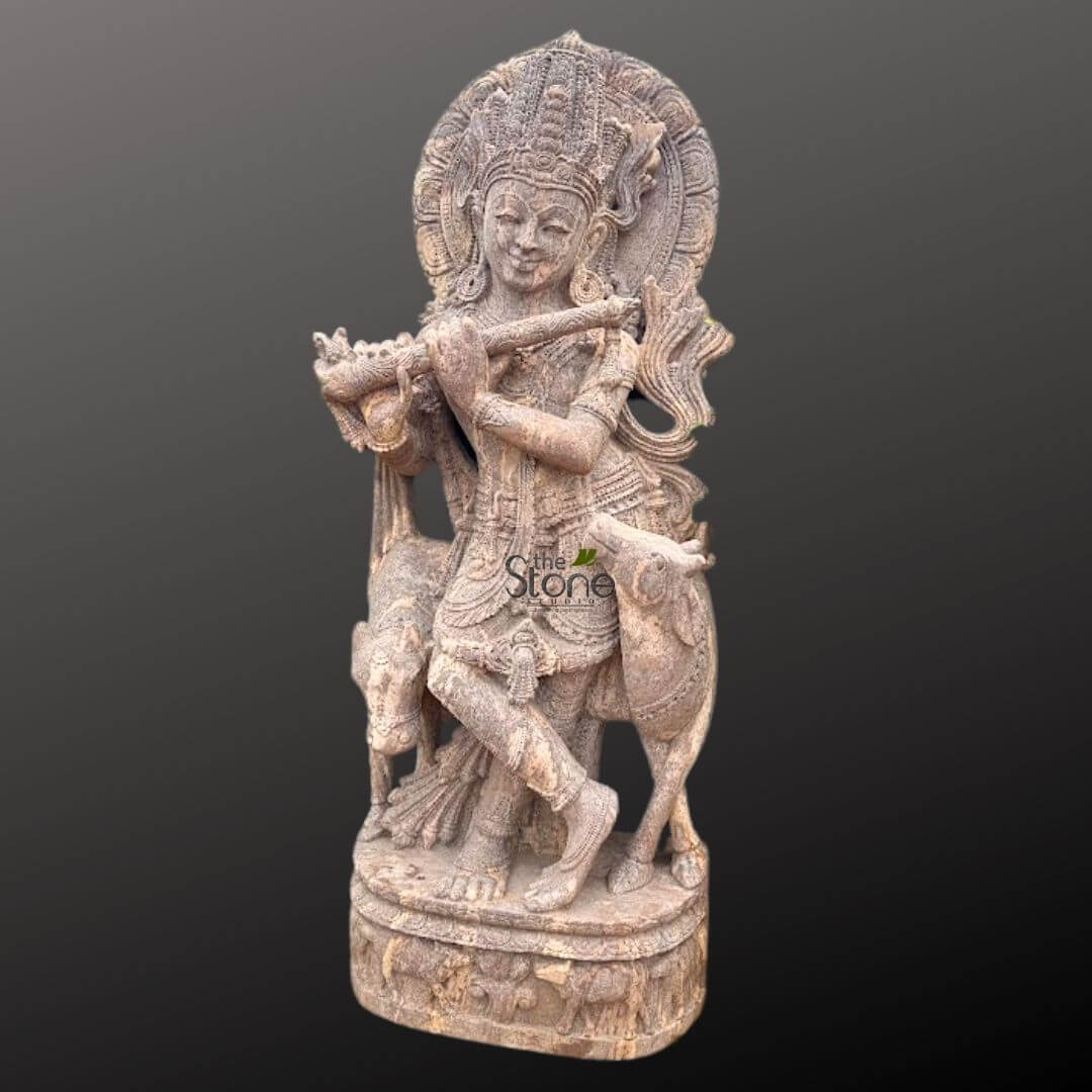 Gopala Krishna statue 5ft 5 meticulously carved, full-length stone sculpture depicts Lord Krishna standing gracefully in a tribhanga (S-shaped) pose, playing his bansuri (flute) held across his chest. The deity wears an elaborate, tiered crown with intricate relief work and has a serene, gentle smile on his face, conveying a sense of divine tranquility and joy. His long, wavy hair flows down his back from beneath the crown. Krishna is adorned with rich ornamentation, including multiple necklaces, armlets, bracelets, and anklets, all exquisitely detailed, suggesting the fine craftsmanship of the piece. He is dressed in traditional flowing garments, a dhoti-like lower garment, and sash, with folds and textures beautifully rendered in the stone. Flanking Krishna on either side are two intricately carved calves or young cows, depicted with realistic features and a gentle posture, looking up towards him, emphasizing his role as a cowherd (Gopala). Behind Krishna's head, a large, circular, ornate halo or prabhamandal is carved, further signifying his divine status and radiating a sense of sacred energy. The entire sculpture rests on a multi-tiered, circular pedestal base, which is also embellished with subtle relief carvings of figures, perhaps devotees or additional animals. The material of the statue appears to be a light brown or sandy-toned natural stone, possibly sandstone, exhibiting a slightly weathered or aged texture that enhances the antique aesthetic and highlights the depth of the carving. The background is a plain, dark gray, making the detailed craftsmanship and the earthy tones of the stone sculpture stand out prominently. This devotional artwork is a prime example of traditional Indian stone carving, representing themes of divine love, music, and nature, making it highly relevant for articles on Hindu mythology, spiritual art, traditional Indian craftsmanship, or decorative garden sculptures.