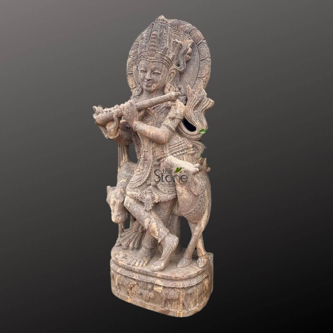 Gopala Krishna statue 5ft 2 highly detailed, handcrafted stone sculpture depicting Lord Krishna, standing in a graceful Tribhanga pose while playing a Bansuri flute. The entire statue, carved from a light brown or earthy beige natural stone, showcases intricate craftsmanship and a slightly textured, rustic finish. Lord Krishna is adorned with an elaborate, multi-tiered crown, intricate necklaces, armbands, and a beautifully draped dhoti (waistcloth) with visible folds and decorative patterns. His face bears a serene, gentle smile, with finely carved features including half-closed eyes that convey a sense of peaceful devotion. Behind his head, a circular halo or 'prabhamandala' is intricately carved with radiating designs, adding to his divine aura. Flanking Krishna are two calves or young cows, sensitively rendered, one standing by his side and the other partially visible, reinforcing his identity as the divine cowherd, Gopala. The sculpture stands on a multi-tiered, rectangular base that is also intricately carved with smaller figures, possibly devotees, or delicate floral and geometric motifs around its perimeter. The statue's overall composition exudes spiritual tranquility and artistic elegance, highlighting traditional Indian stone carving techniques. The "the Stone" watermark is visible on the lower front of the statue, likely indicating the artisan or seller. This piece would be suitable for a temple, home altar, garden, or as a significant decorative art piece for cultural and spiritual enthusiasts, connecting to themes of Hindu mythology, ancient Indian art, and artisanal stone carving.
