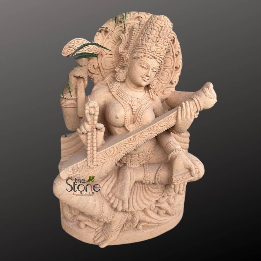 Saraswati Maa Statue 2.5ft 3 beautifully detailed and serene stone sculpture of the Hindu Goddess Saraswati, crafted from a light beige or sandstone-colored material, stands prominently against a plain dark gray background. The deity is depicted in a graceful, seated posture with four arms (Chaturbhuja), symbolizing her divine power and multifaceted nature. Her two primary hands delicately hold and play a intricately carved veena, a traditional Indian string instrument, with her fingers positioned as if plucking the strings. Her upper right hand gently holds a japa mala (rosary beads), representing her association with meditation, spirituality, and the pursuit of knowledge. From her upper left hand, which appears to be holding a kalasha (water pot) or a similar vessel, vibrant green foliage, possibly a snake plant or a similar hardy plant, gracefully emerges upwards, indicating its potential use as a unique planter or a garden feature. This integration of living plant life suggests a connection to nature, growth, and vitality, making it suitable for both indoor spiritual spaces and outdoor garden settings. Saraswati's face exudes tranquility and wisdom, with finely sculpted features, downcast eyes, and a gentle, peaceful smile. She is elaborately adorned with a tall, ornate crown (kiritamukuta), multiple layered necklaces, prominent earrings, bangles on her wrists, and anklets. Her traditional drapery and the base she sits upon are also meticulously carved with intricate patterns and folds, showcasing exceptional artistry and craftsmanship. Behind her head, a circular, carved halo or prabhamandala with radiating patterns signifies her divine aura and spiritual significance. The sculpture's textured, matte finish highlights the natural beauty of the stone, lending an earthy and ancient feel. The overall presentation emphasizes its value as a piece of devotional art, a spiritual home decor item, or an elegant garden sculpture. A subtle "the Stone Studio" watermark is visible in the lower left, identifying the creator or vendor of this exquisite handcrafted piece, which would naturally be featured in articles about traditional Indian sculpture, Hindu religious art, spiritual home design, bespoke stone carvings, or unique garden planters.