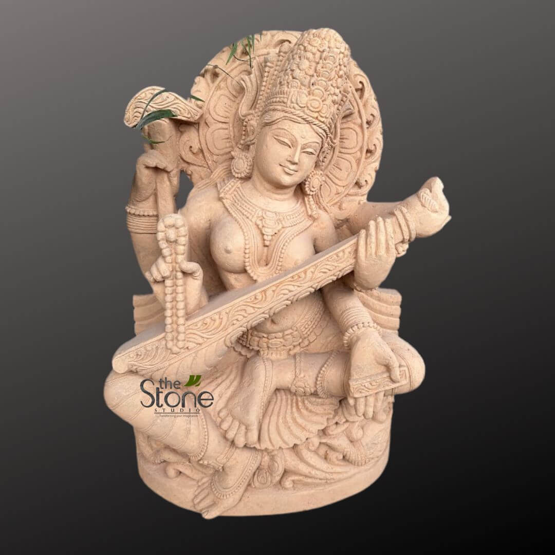 Saraswati Maa Statue 2.5ft 2 beautifully detailed and grand stone sculpture depicts the Hindu Goddess Saraswati, the revered deity of knowledge, music, arts, wisdom, and learning. She is gracefully seated in a traditional meditative posture, her legs folded, while delicately playing the veena, a classical Indian stringed instrument, which rests diagonally across her lap. Her serene face features gently downcast eyes, a soft, peaceful smile, and is crowned with an intricately carved, multi-tiered mukuta (headdress) that showcases fine floral and geometric patterns. Saraswati is depicted with four arms, a common attribute of Hindu deities, symbolizing her omnipresence and power. Her primary two hands are engaged with the veena, while one of her back hands holds a mala (rosary beads), and the other appears to grasp a sacred palm leaf manuscript or a lotus bud, with some real green foliage subtly placed near it for natural presentation. Every element of the sculpture showcases meticulous craftsmanship, from her ornate necklaces, armlets, bracelets, and anklets to the flowing drapery of her garments, which are adorned with delicate patterns and folds, highlighting the contours of her form. Behind her head, a detailed circular prabhamandala (aura or halo) with radiating patterns further enhances her divine presence. The sculpture is crafted from a light beige or reddish-brown natural stone, likely sandstone, exhibiting a matte, slightly granular texture that beautifully highlights the depth and precision of the carving work. The base on which she sits is also intricately carved, providing a sturdy and aesthetically pleasing foundation. This exquisite piece exemplifies the artistry and dedication of "The Stone Studio," whose distinct logo is subtly visible at the lower left base of the sculpture. The intricate details, the expressive serenity of the goddess, and the natural stone material make this a magnificent example of traditional Indian stone carving, perfect for enhancing a spiritual space, a contemplative garden, or as a focal point in a culturally inspired interior design, embodying themes of peace, wisdom, and artistic expression.