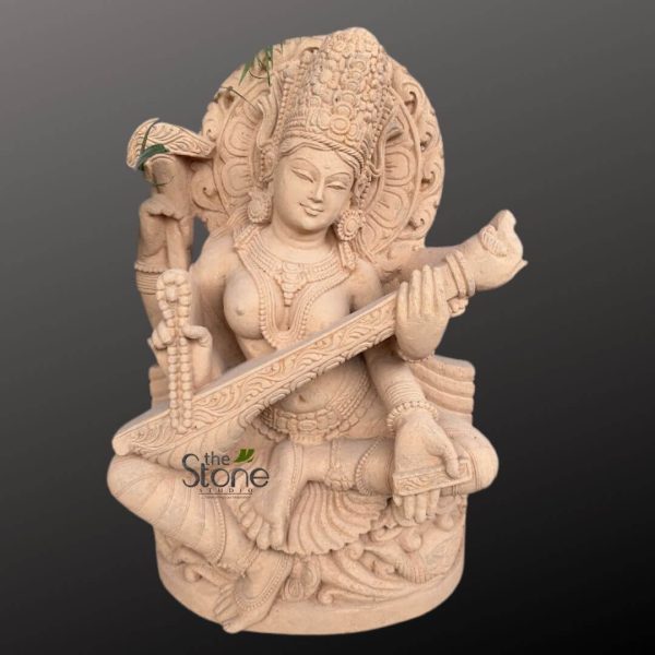 beautifully carved, light beige sandstone statue of the Hindu Goddess Saraswati, seated gracefully in a meditative posture. Her serene face, with gently smiling lips and downcast eyes, conveys peace and wisdom. She is adorned with an elaborate crown (Kiritamukuta) featuring intricate floral and geometric patterns, and a circular, ornate prabhamandala (halo) frames her head, symbolizing divine aura. Saraswati is depicted with four arms, a common representation in Hindu iconography. Her primary two hands delicately hold a richly detailed veena (a stringed musical instrument, similar to a lute), its neck extending upwards and its body resting across her lap. The veena itself is embellished with carved designs. Her upper right hand holds a rosary (mala), signifying spirituality and meditation, while her upper left hand clutches a palm-leaf manuscript (Pustaka), representing knowledge, learning, and the scriptures. The goddess wears finely sculpted jewelry, including multiple necklaces, armlets, bangles on her wrists, and anklets on her feet, all carved directly into the stone. Her drapery, a flowing sari or dhoti, is also intricately detailed with folds and patterns, suggesting soft fabric despite being rigid stone. A small, delicate green plant or vine appears to sprout from behind her crown, adding a natural, organic touch to the ancient craft. She is seated on a decorative, layered pedestal that features carved leaf and scroll motifs, complementing the divine figure. The statue’s surface exhibits the natural, slightly textured quality of sandstone, giving it an earthy yet refined appearance. The entire sculpture is presented against a plain, dark grey background, making the warm, monochromatic tones of the sandstone stand out prominently. A subtle "the Stone Studio" logo is visible at the bottom left. This detailed piece exemplifies traditional Indian stone carving, embodying the essence of art, knowledge, and spiritual devotion, making it a powerful statement for spiritual home decor, garden art, or cultural collections.