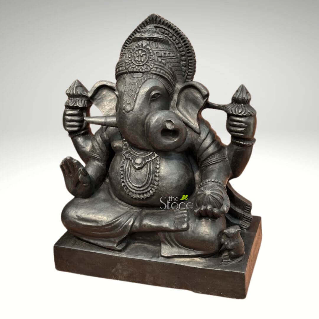 Small Ganesha statue for Home 15" 5 meticulously carved, dark grey stone statue of Lord Ganesha, the revered Hindu elephant-headed deity, is presented centrally against a plain, light grey background. Ganesha is depicted seated gracefully in a meditative posture, likely padmasana (lotus pose), on a solid, rectangular dark grey plinth. The sculpture exhibits a rich, deep charcoal grey or black hue, suggesting polished natural stone like granite or basalt, with subtle highlights that emphasize its intricate details and robust form. Ganesha's iconic elephant head features large, expressive ears, a gently curving trunk, and one broken tusk – a common iconography symbolizing wisdom and sacrifice, often associated with his role as the scribe of the Mahabharata. His eyes are half-closed, conveying a sense of profound serenity and contemplation. He wears an elaborately sculpted crown or kirīṭamakuṭa adorned with traditional Indian patterns and a decorative forehead band. His portly belly, a symbol of abundance and prosperity, is prominent, and he is adorned with multiple intricate necklaces, bangles on his wrists, and a sacred thread (yagnopaveetam) draped across his chest. The folds of his draped garment are visible around his legs. Ganesha is depicted with four arms, holding his traditional attributes: in his upper right hand, he grasps an ankusha (goad), and in his upper left hand, he holds a pasha (noose). His lower right hand is raised in abhaya mudra, a gesture of blessing and dispelling fear. His lower left hand gently holds a bowl filled with modaks (sweet dumplings), symbolizing rewards and joy. At his lower right foot, a small, alert mouse (mooshak or akhuket), Ganesha's traditional vehicle, sits attentively, looking towards the deity and completing this iconic representation. The overall craftsmanship highlights traditional Indian artistry, making this stone idol a striking piece of spiritual decor or religious art, ideal for discussions on Hindu deities, stone carving, or the significance of Ganesha in spiritual practices.