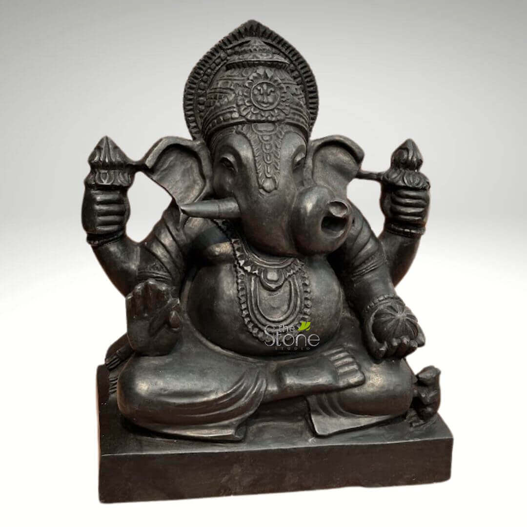 Small Ganesha statue for Home 15" 6 meticulously crafted, dark charcoal grey stone statue of Lord Ganesha is centered in the frame, depicted in a traditional meditative pose. The Hindu deity, revered as the remover of obstacles, god of wisdom, and harbinger of prosperity, sits cross-legged on a solid, rectangular base. His iconic elephant head features large, gracefully fanned ears and a trunk that curves gently to his left, reaching towards the modak in his lower left hand. One tusk, typically his left, is broken, symbolizing his sacrifice for knowledge and dedication to writing the Mahabharata. Ganesha wears an elaborate, tiered crown adorned with intricate floral and geometric patterns, topped by a textured crest and featuring a prominent central medallion. His forehead bears a spiritual mark, enhancing his serene expression. He is depicted with four arms, each holding symbolic attributes: his upper right hand grasps a finely detailed ankusha (goad), while his upper left holds a pasha (noose). His lower right hand is raised in the benevolent Abhaya Mudra, offering blessings and protection, and his lower left hand delicately holds a spherical modak (sweet dumpling), representing rewards and spiritual fulfillment. The statue showcases Ganesha's characteristic portly belly, symbolizing abundance and generosity, adorned with an intricately carved, multi-layered necklace featuring a central pendant, along with decorative armlets and bangles. At the base, to his lower right, sits his loyal vahan (mount), Mushika the mouse, attentively looking up at him. The stone has a smooth, matte finish that highlights the intricate details and expert craftsmanship of the carving. The statue is evenly lit against a soft, neutral grey background, which emphasizes its rich, dark hue and detailed textures. This exquisite Ganesha idol serves as a powerful symbol of good fortune and auspicious beginnings, making it a perfect piece for spiritual decor, a home altar, or as a timeless representation of traditional Indian religious art.