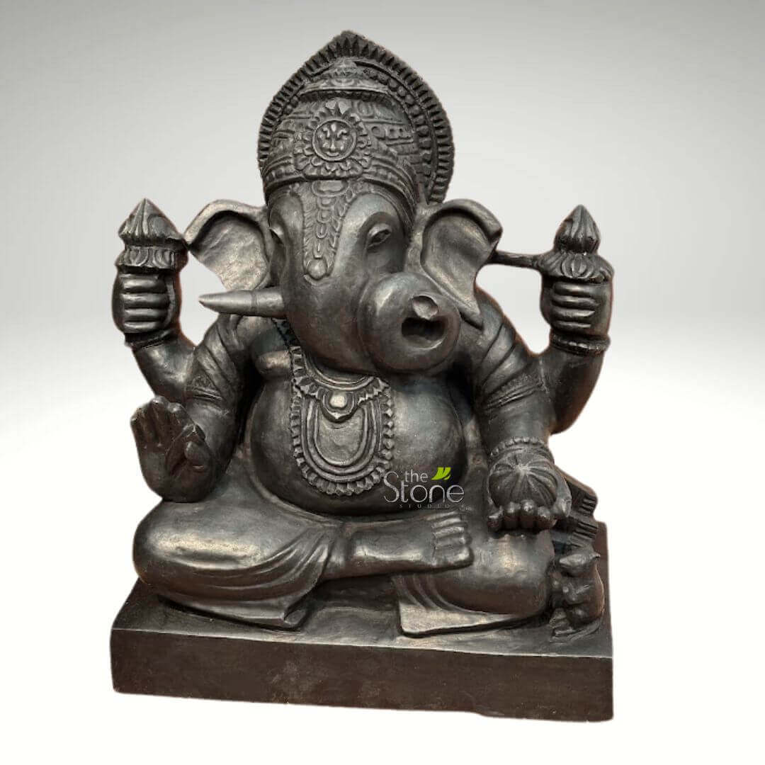 Small Ganesha statue for Home 15" 4 striking and detailed dark gray or black stone sculpture of the Hindu deity Ganesha, seated in a meditative cross-legged lotus posture (Padmasana) on a simple, rectangular plinth. Ganesha, known as the remover of obstacles and god of wisdom and prosperity, is depicted with his iconic elephant head, featuring large, gracefully curved ears, a single tusk, and a long, curled trunk that gently reaches towards a round sweet, likely a modak, held in his lower left hand. His body is portrayed with a traditional pot-belly, signifying abundance. He has four arms, a common representation for Ganesha. In his upper right and left hands, he holds slender, pointed objects resembling lotus buds or ritual implements, while his lower right hand is raised in the Abhaya mudra, a gesture of blessing and fearlessness, with the palm facing outwards. The sculpture is rich in intricate details, showcasing finely carved adornments. Ganesha wears an elaborate crown or headdress with a halo-like element behind it, along with a prominent, multi-layered necklace, bracelets, and anklets. The folds of his draped lower garment are also meticulously rendered. A small, intricately carved mouse, his vahana (mount), is visible seated at his lower right side, near his knee. The material appears to be a polished dark stone, possibly granite or a similar stone, giving the idol a profound and elegant appearance. The surface has a smooth, almost metallic sheen that highlights the craftsmanship and depth of the carving. The sculpture stands against a clean, uniformly light gray background, allowing all its features to be clearly visible. A discreet watermark logo "the Stone Studio" is visible in the lower right, indicating the artisan or source. This majestic Ganesha idol would serve as a powerful and beautiful centerpiece for spiritual home decor, temple art, or as a symbol of cultural heritage.