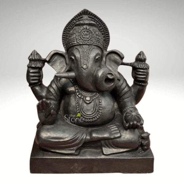 meticulously carved dark gray or black stone statue of the Hindu deity Ganesha is presented centrally against a plain, light gray background, highlighting its intricate details. The four-armed, elephant-headed god is depicted in a serene, meditative seated pose, specifically a form of Padmasana or Lalitasana, atop a simple rectangular base. Ganesha's large, fan-shaped ears frame his gentle face, featuring a prominent, single tusk on his right side (viewer's left), signifying his Ekadanta form and connection to wisdom and sacrifice. His trunk gracefully curls to his left (viewer's right), reaching towards a modak (sweet dumpling) held in his lower left hand. His upper right hand (viewer's left) firmly grasps an ankusha (goad), symbolizing control over desires and obstacles. His upper left hand (viewer's right) holds a pasha (noose), representing attachment and restraint, while his lower right hand (viewer's left) is raised in abhaya mudra, offering blessing and dispelling fear. The deity's characteristic potbelly (Lambodara) is pronounced, adorned with a multi-strand necklace and a sacred thread (upanayana) draped diagonally across his chest. He wears an elaborate, tiered crown (mukuta) with intricately carved floral and geometric patterns, along with armbands and bracelets, all rendered with fine precision in the dark stone. At the base, near Ganesha's right foot, his traditional vehicle (vahana), a tiny, alert mouse (Mushika), is subtly carved, adding another layer of traditional iconography. The stone material, with its slightly matte finish, gives the statue a weighty and timeless feel, emphasizing the craftsmanship involved. This detailed sculpture, likely from "The Stone Studio" as indicated by the watermark, is an excellent representation for articles exploring Hindu mythology, devotional art, traditional Indian sculpture, or as a symbol of wisdom, prosperity, and the removal of obstacles.