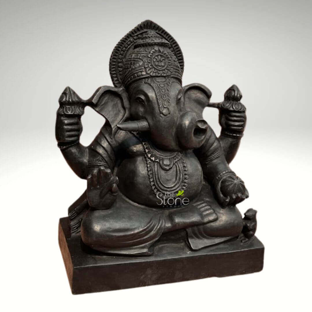 Small Ganesha statue for Home 15" 3 majestic and intricately carved dark stone sculpture of Lord Ganesha, the elephant-headed Hindu deity, is presented front-on against a soft, light grey background. The statue, likely crafted from black granite or a similar dark, heavy stone, exudes a profound sense of serenity and strength, with a rich, matte finish that accentuates its detailed artistry. Ganesha is depicted seated in a cross-legged meditative posture on a simple, sturdy rectangular plinth. His iconic elephant head features large, fanned ears and an ornate, multi-layered crown with intricate traditional patterns and a prominent central motif. His serene face shows eyes cast downward in contemplation, while his trunk curls gracefully to his left, appearing to reach towards a sweet treat (modak) held in one of his lower hands. One tusk is notably broken, a classic iconographic detail associated with Ganesha. He is portrayed with four arms, each holding symbolic objects. His upper right hand holds an 'ankus' (goad), signifying spiritual guidance and the removal of obstacles. His upper left hand grasps a 'pasha' (noose), representing the control of worldly attachments. His lower right hand is raised in the 'Abhaya Mudra,' a gesture of reassurance and fearlessness, while his lower left hand cradles what appears to be a 'modak' or spherical sweet, symbolizing rewards and spiritual liberation. Ganesha's corpulent pot belly, a symbol of prosperity and generosity, is prominent. He is adorned with layered necklaces or malas, armlets, and wristlets, all rendered in the same dark stone, contributing to the statue's cohesive and elegant aesthetic. Draped fabric is sculpted around his lower body, indicating his traditional attire. A tiny, well-defined mouse, Ganesha's 'vahana' or vehicle, is subtly carved beside his right foot on the base, completing the traditional representation. The watermark "The Stone Studio" is visible on the lower front, suggesting the origin or craftsmanship of this beautiful Ganesha idol, suitable for spiritual reflection, home décor, or as a collector's item embodying wisdom, prosperity, and auspicious new beginnings.