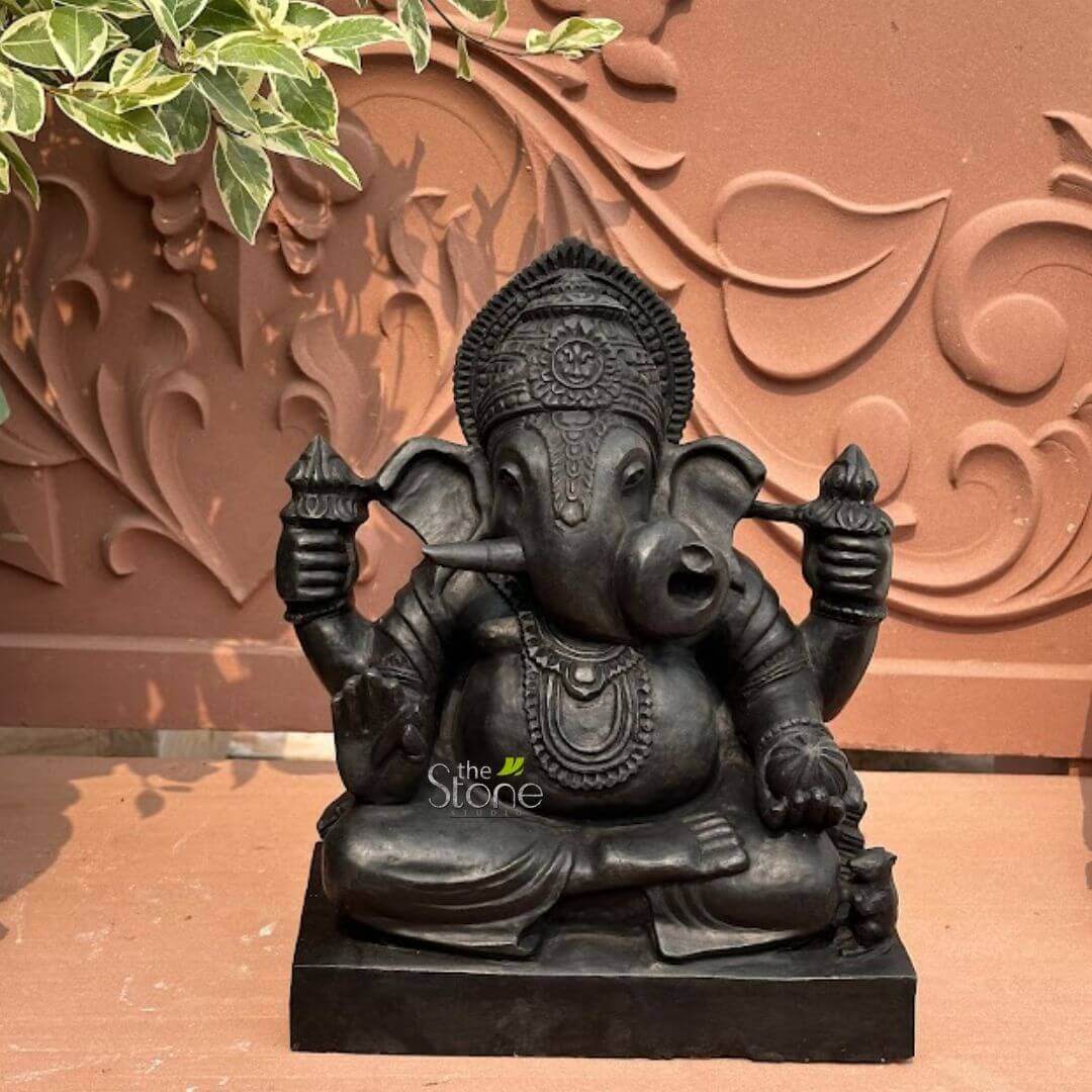 Small Ganesha statue for Home 15" 2 meticulously crafted, dark charcoal-grey stone sculpture of the Hindu deity Ganesha, depicted in a serene, seated posture, possibly Padmasana, on a simple rectangular plinth. The elephant-headed god is rendered with intricate details, showcasing his large, fanned ears, expressive eyes, and a gracefully curled trunk reaching towards a spherical sweet delicacy (modak) cradled in his lower left hand. His lower right hand distinctively holds his iconic broken tusk, symbolizing sacrifice and the pursuit of knowledge and wisdom. Ganesha is adorned with an elaborately carved crown featuring a central motif, multiple layered necklaces, detailed armbands, and a prominent sacred thread (yajnopavita) draped across his robust physique, all carved into the dark stone. The dark, almost black, stone of the Ganesha idol stands out against a textured reddish-brown background, which appears to be a terracotta or carved sandstone panel. This background features elegant, flowing floral or vine-like motifs in relief, hinting at traditional Indian architectural or decorative design. In the upper left corner of the image, vibrant green leaves with variegated white edges from a plant gently cascade downwards, adding a natural, organic touch and a contrasting splash of color to the earthy tones of the setting. The entire scene rests on a flat, reddish-brown surface, creating a cohesive and spiritually resonant ambiance. This detailed Ganesha murti, with its rich texture and profound symbolism, suggests a piece intended for spiritual contemplation, an exquisite item of Indian home decor, or a representation of fine stone craftsmanship. A subtle "the Stone Studio" watermark is visible on the lower front of the statue, indicating its origin or creator.