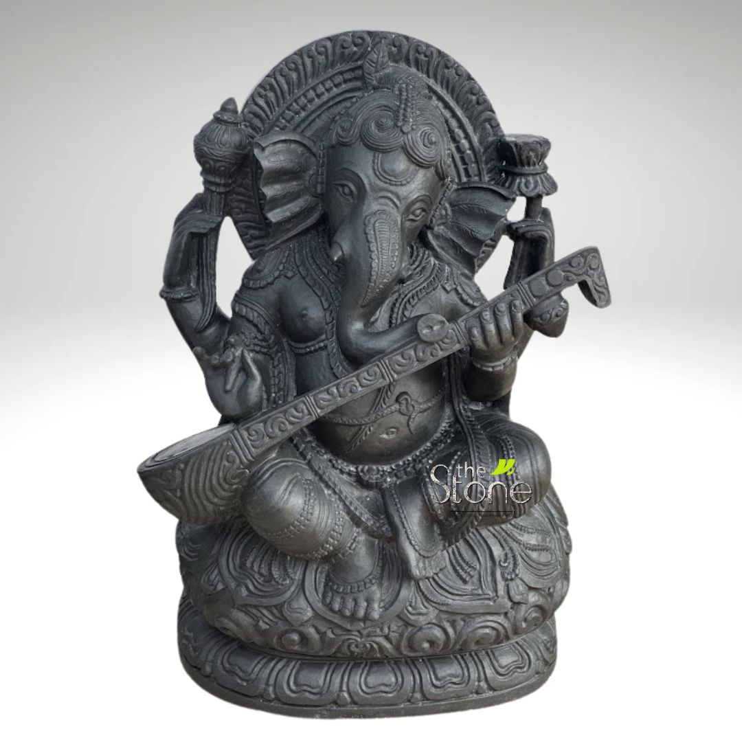 Veena Ganesha Statue 2ft 4 This is a detailed, dark gray stone statue of the Hindu deity Ganesha, depicted seated gracefully and playing a musical instrument. The sculpture showcases Ganesha in a cross-legged, meditative posture (Padmasana) on an intricately carved lotus base. His iconic elephant head features large, fanned ears, a gently curving trunk turned towards his left, and expressive, small eyes. A detailed, multi-tiered crown adorns his head, and a decorative aureole (prabhamandala) carved with traditional patterns frames his upper body from behind, adding to the grandeur. Ganesha is portrayed with four arms, symbolizing his divine power. His two lower, front hands are positioned to delicately hold and play a veena, a traditional Indian stringed instrument, which rests across his lap. The veena itself is beautifully rendered with carved patterns along its body and neck. In his upper left hand, Ganesha holds a small, round modaka (a sweet delicacy), while his upper right hand clasps a lotus bud or flower. The deity is richly adorned with numerous finely carved ornaments, including elaborate necklaces, armlets, bracelets, anklets, and a sacred thread (yagnopavitam) draped across his robust belly, a characteristic feature. The entire statue appears to be crafted from a heavy, dark gray or black stone, such as granite or soapstone, presenting a matte finish that accentuates the intricate details and textural contrasts of the carving. The light, plain background with a subtle gradient ensures that the focus remains entirely on this magnificent and spiritual piece of art. This Ganesha with Veena idol beautifully represents Ganesha as the Lord of Arts, Music, Wisdom, and the Remover of Obstacles, making it a significant piece for spiritual contemplation, home decor, or as a symbol of cultural heritage and artistic expression.