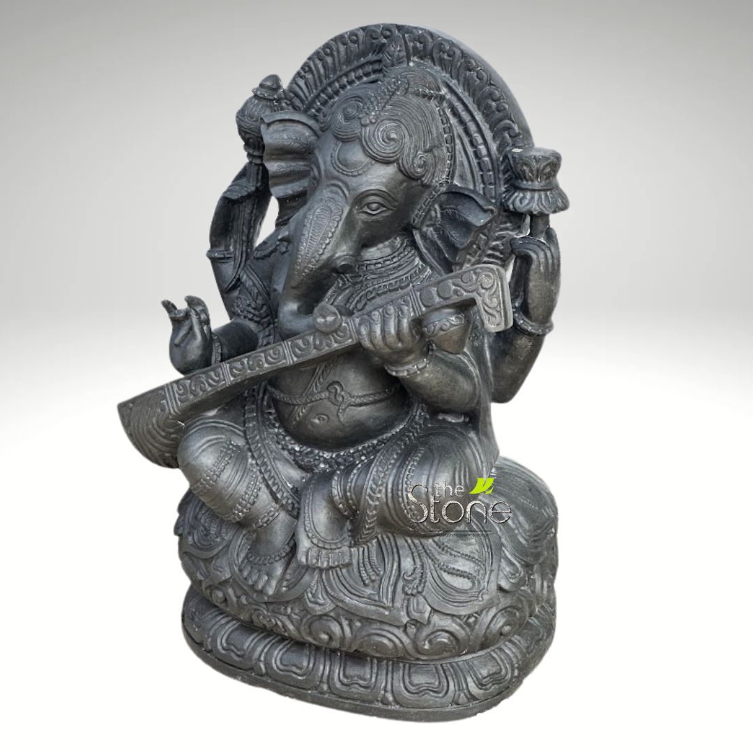 Veena Ganesha Statue 2ft 3 This alt text describes a finely detailed dark gray or black stone sculpture of Lord Ganesha, depicted in a serene and artistic pose. The elephant-headed Hindu deity is shown seated in a meditative, cross-legged posture, likely padmasana, upon an elaborate double-tiered lotus pedestal (padma peetha). Ganesha's characteristic features include his large, gently flared ears, intelligent eyes, and a gracefully curved trunk, which sweeps towards his left side. He is portrayed with four arms, a common depiction for Ganesha, symbolizing his divine power and attributes. His two main hands are delicately positioned to hold and play the Veena, an ancient Indian stringed musical instrument, across his lap. The details of the instrument, from its long neck to the intricate carvings on its body, are clearly visible. One of his upper right hands holds a small, auspicious lotus bud or decorative flourish, while his upper left hand is raised in a gesture of blessing or fearlessness (Abhaya Mudra), conveying peace and protection. The entire sculpture is rendered with exquisite craftsmanship, showcasing intricate details. Ganesha wears an ornate crown (mukuta) and a halo-like arch behind his head, both adorned with traditional patterns. His body is decorated with various pieces of jewelry, including necklaces, armlets, anklets, and a sacred thread (yagnopavita). His dhoti (lower garment) and sash are also sculpted with fine textures and patterns. The dark, charcoal-gray, or black stone material (possibly granite or a similar dark stone) lends the sculpture a timeless, dignified, and matte finish. The subtle interplay of light and shadow emphasizes the three-dimensional depth and intricate carvings of the statue. The background is a plain, diffused light gray, allowing the entire focus to remain on the detailed artistry of the Ganesha idol. This visually rich sculpture, embodying Ganesha as the patron of arts, wisdom, and music, would be an excellent visual for articles discussing Hindu iconography, traditional Indian stone carving, spiritual home decor, the significance of the Veena, or the cultural importance of Lord Ganesha as the remover of obstacles and god of new beginnings.