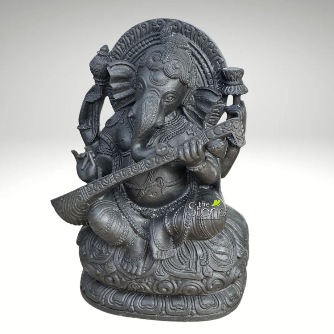 Veena Ganesha Statue 2ft 2 striking, full-length image of an intricately detailed dark gray Ganesha statue, depicted in a serene, seated posture playing a veena. This Hindu deity, revered as the god of wisdom, intellect, arts, and the remover of obstacles, is rendered with exceptional craftsmanship, appearing to be carved from dark stone or a high-quality polyresin with an antique or metallic charcoal finish. Ganesha's iconic elephant head is prominent, featuring large, gracefully fanned ears, expressive eyes, and a gently curved trunk. His four arms are elegantly positioned: the primary two hands delicately hold and play a long, elaborately carved veena, a traditional Indian stringed instrument, showcasing Ganesha's connection to music and the arts. His upper left hand is raised in the 'Abhaya Mudra,' a gesture of reassurance and fearlessness, while his upper right hand holds a small, symbolic item, possibly a modak (sweet dumpling) or a lotus bud, representing spiritual purity. The deity is adorned with rich, textural details including multiple necklaces, armlets, and bracelets, all intricately sculpted to reflect traditional Indian jewelry. His dhoti (garment) is also embellished with fine, traditional patterns. Ganesha sits comfortably cross-legged (in a lotus-like posture) upon a multi-layered, ornate lotus pedestal (Padmasana base), with each petal meticulously defined and carved. Behind his head, an exquisite, circular 'prabhamandala' or aura is carved with radiating, intricate patterns, framing the deity and enhancing its majestic and divine presence. The entire sculpture is presented in a monochrome dark gray tone, which accentuates the deep textures, shadows, and fine details created by the carving work. The plain, soft gray to white gradient background ensures that the Ganesha idol stands out clearly, emphasizing its artistic and spiritual significance. This highly decorative murti exemplifies a blend of spiritual symbolism and artistic excellence, making it an ideal focal point for spiritual decor, home altars, garden settings, or as a meaningful symbol of knowledge, success, and positive new ventures.