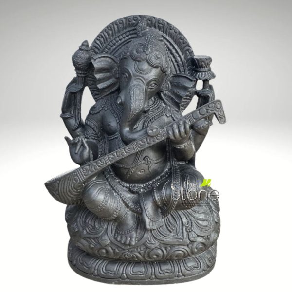 striking and intricately detailed dark grey stone statue depicting Lord Ganesha, the revered Hindu deity known as the remover of obstacles and patron of arts and sciences. The elephant-headed god is shown in a serene, seated posture, likely Padmasana or lotus pose, on a richly carved multi-layered lotus pedestal. Ganesha holds a beautifully crafted veena, a traditional Indian stringed musical instrument, across his lap, with his lower two hands positioned as if playing it, symbolizing his connection to music, art, and knowledge. His upper left hand holds a shankha (conch shell), while his upper right hand holds what appears to be a lotus bud or an offering bowl. The statue's surface boasts a matte, textured finish, characteristic of natural stone like dark granite or charcoal sandstone, enhancing the depth and detail of the carving. Every element, from Ganesha's elaborate crown and large ears to the delicate ornamentation on his clothing and the prabhavali (aura/arch) behind him, is meticulously rendered with traditional motifs. The clean, light background ensures the focus remains entirely on this exquisite piece of spiritual art or devotional home decor. This Ganesha idol playing veena perfectly illustrates themes of wisdom, creativity, and divine blessings, making it ideal for articles discussing Hindu iconography, Indian craftsmanship, spiritual well-being, or traditional decor.