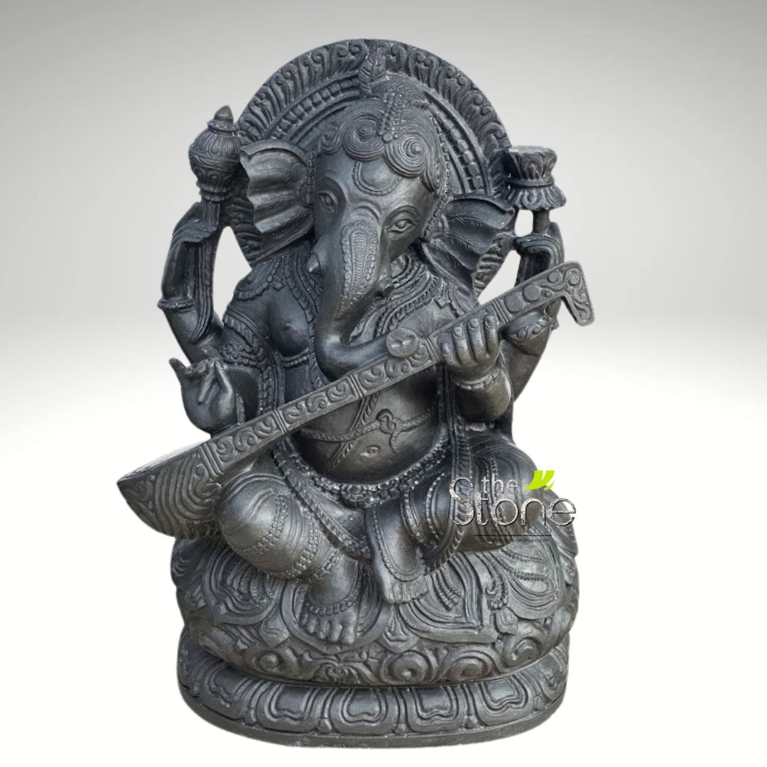 Veena Ganesha Statue 2ft 1 striking and intricately detailed dark grey stone statue depicting Lord Ganesha, the revered Hindu deity known as the remover of obstacles and patron of arts and sciences. The elephant-headed god is shown in a serene, seated posture, likely Padmasana or lotus pose, on a richly carved multi-layered lotus pedestal. Ganesha holds a beautifully crafted veena, a traditional Indian stringed musical instrument, across his lap, with his lower two hands positioned as if playing it, symbolizing his connection to music, art, and knowledge. His upper left hand holds a shankha (conch shell), while his upper right hand holds what appears to be a lotus bud or an offering bowl. The statue's surface boasts a matte, textured finish, characteristic of natural stone like dark granite or charcoal sandstone, enhancing the depth and detail of the carving. Every element, from Ganesha's elaborate crown and large ears to the delicate ornamentation on his clothing and the prabhavali (aura/arch) behind him, is meticulously rendered with traditional motifs. The clean, light background ensures the focus remains entirely on this exquisite piece of spiritual art or devotional home decor. This Ganesha idol playing veena perfectly illustrates themes of wisdom, creativity, and divine blessings, making it ideal for articles discussing Hindu iconography, Indian craftsmanship, spiritual well-being, or traditional decor.