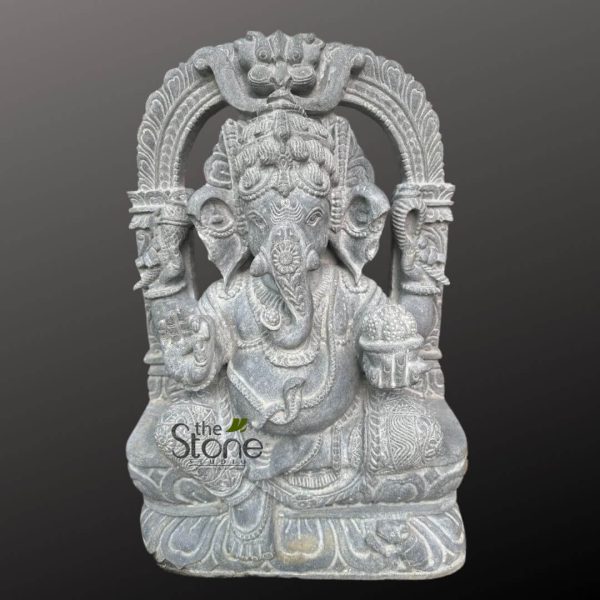 beautifully detailed and intricately carved grey stone statue of Lord Ganesha is presented centrally against a plain, solid dark grey background, highlighting its exquisite craftsmanship. The revered Hindu deity is depicted in a traditional seated posture, likely Padmasana or Virasana, on a stylized lotus pedestal, symbolizing purity and spiritual awakening. Ganesha features his iconic elephant head with large, fanned ears and small, deep-set eyes, conveying wisdom. His trunk elegantly curls towards his left (viewer's right), touching a modak (sweet dumpling) held in his lower right hand, which is often symbolic of rewards for spiritual pursuits. An elaborate, multi-tiered crown or mukuta adorns his head, meticulously carved with fine patterns and a prominent tilak mark on his forehead. He is depicted with four arms, a common iconography for Ganesha. His upper left hand holds an ankusha (elephant goad), symbolizing control over desires, while his upper right hand grasps a pasha (noose), representing the binding nature of worldly attachments. His lower left hand is raised in abhaya mudra, a gesture of fearlessness and blessing, offering protection to devotees. The statue showcases Ganesha's characteristic pot-belly, symbolizing prosperity and the ability to consume all sorrows. He is adorned with numerous carved ornaments, including necklaces, armlets, anklets, and a sacred thread (yagnopavita) draped across his body. His dhoti (waistcloth) is also richly detailed with ornate patterns. Behind the deity, a highly ornamental prabhamandala (aura or arch) frames the figure, extending upwards and outwards. This decorative arch features classical South Indian temple motifs, including stylized makara (mythical aquatic creatures) at the base of the pillars and what appears to be a kirtimukha (glorious face) at the apex, all surrounded by intricate scrollwork, floral designs, and decorative carvings that enhance the statue's grandeur. At the very base, near Ganesha's feet, his vahana, a small mouse (Mooshika), is subtly carved. The statue appears to be crafted from a natural, fine-grained grey stone, possibly granite or a similar type of rock, which allows for the high level of detail and texture seen throughout the piece. The overall impression is one of profound artistry, spiritual significance, and skilled traditional Indian stone carving. A faint watermark for "the Stone Studio" with green leaf accents is visible in the lower-left portion of the image. This stone sculpture is a magnificent example of a religious artifact or a premium piece of artistic home decor.